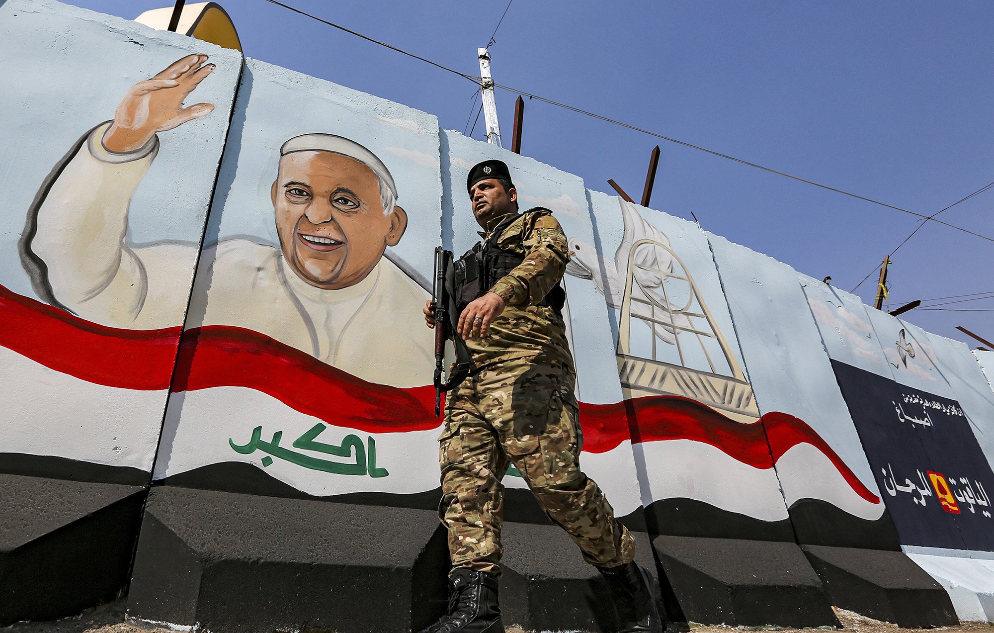 L'image montre un soldat en uniforme, armé et en marche, devant un mur peint. Sur le mur, il y a une fresque représentant un homme souriant et levant la main, probablement symbolisant une figure importante ou un leader. Le fond de la fresque inclut des éléments décoratifs et des couleurs qui évoquent le drapeau irakien. L'environnement semble être une zone urbaine, marquée par des installations de sécurité.