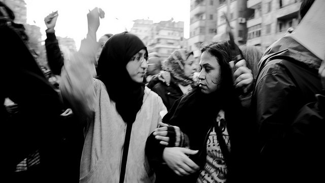 L'image montre un groupe de personnes lors d'une manifestation. Deux jeunes femmes, l'une portant un hijab et l'autre les cheveux détachés, semblent échanger des mots tout en levant les bras, ce qui indique un sentiment d'engagement et de solidarité. L'ambiance est dynamique, et le noir et blanc donne une atmosphère intense à la scène. En arrière-plan, on peut apercevoir d'autres manifestants, créant une impression de mouvement et de rassemblement.