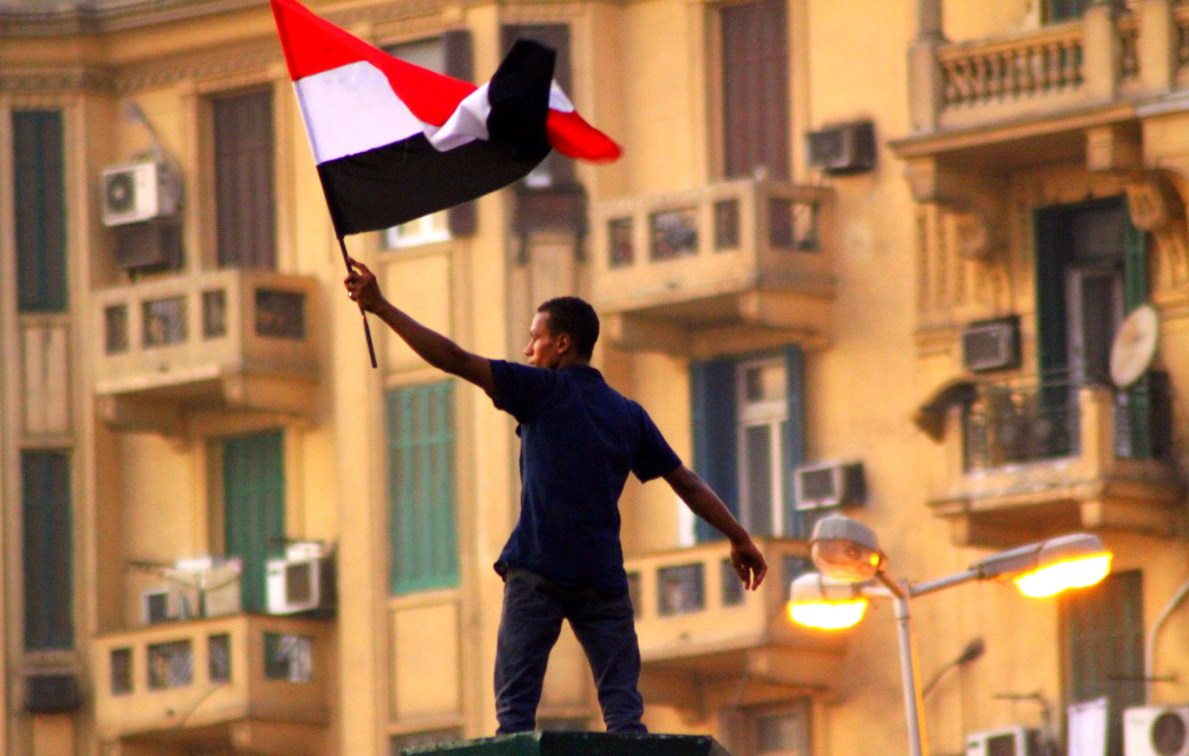 L'image montre un homme brandissant un drapeau noir, blanc et rouge, probablement un drapeau national. Il se tient debout sur une structure, manifestant une posture de célébration ou de protestation. En arrière-plan, on peut apercevoir des bâtiments avec des balcons et des climatiseurs, ce qui suggère un environnement urbain. L'atmosphère semble chargée d'une forte émotion, possiblement liée à un événement politique ou social.