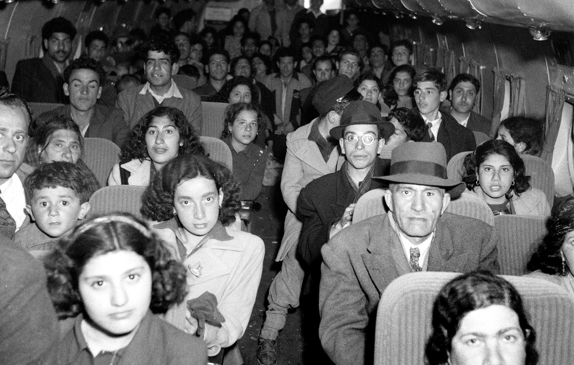 L'image montre l'intérieur d'un avion rempli de passagers. On peut voir un groupe diversifié de personnes, y compris des adultes et des enfants, assis sur des rangées de sièges. Les expressions des passagers varient, certains semblent sérieux, tandis que d'autres regardent droit devant. L'ambiance semble être celle d'un voyage, probablement dans un contexte historique. L'éclairage de l'image est en noir et blanc, ce qui lui donne une atmosphère vintage.