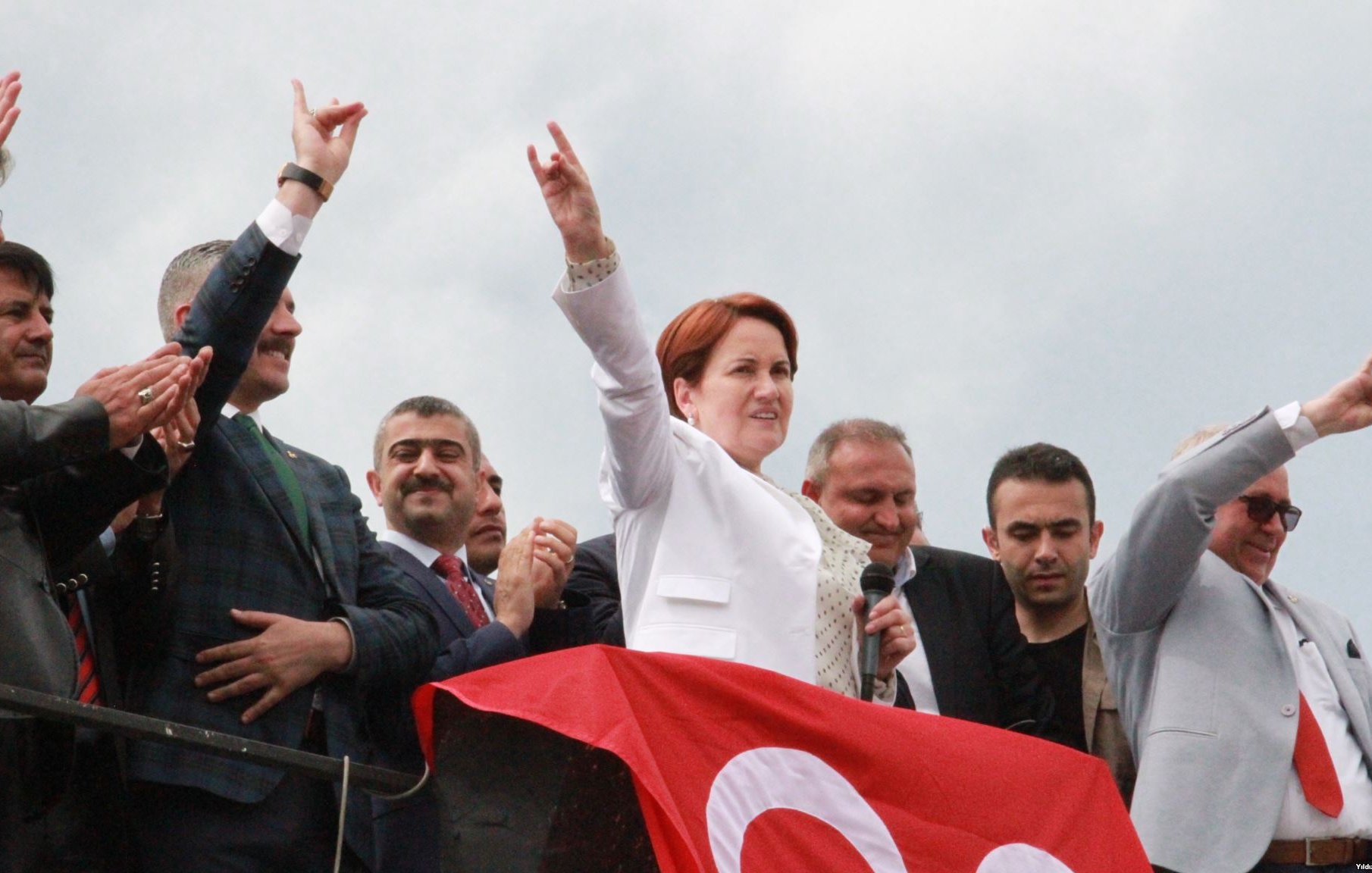 L'image montre un groupe de personnes sur une scène, avec une femme au centre qui lève le bras en signe de victoire tout en tenant un micro. Elle porte une tenue blanche et semble s'adresser à la foule. Derrière elle, plusieurs hommes, dont certains applaudissent, sont visibles. Une grande bannière rouge est accrochée sur la scène, ajoutant une touche de couleur à l'image, avec un ciel nuageux en arrière-plan. L'ambiance semble festive et militante.