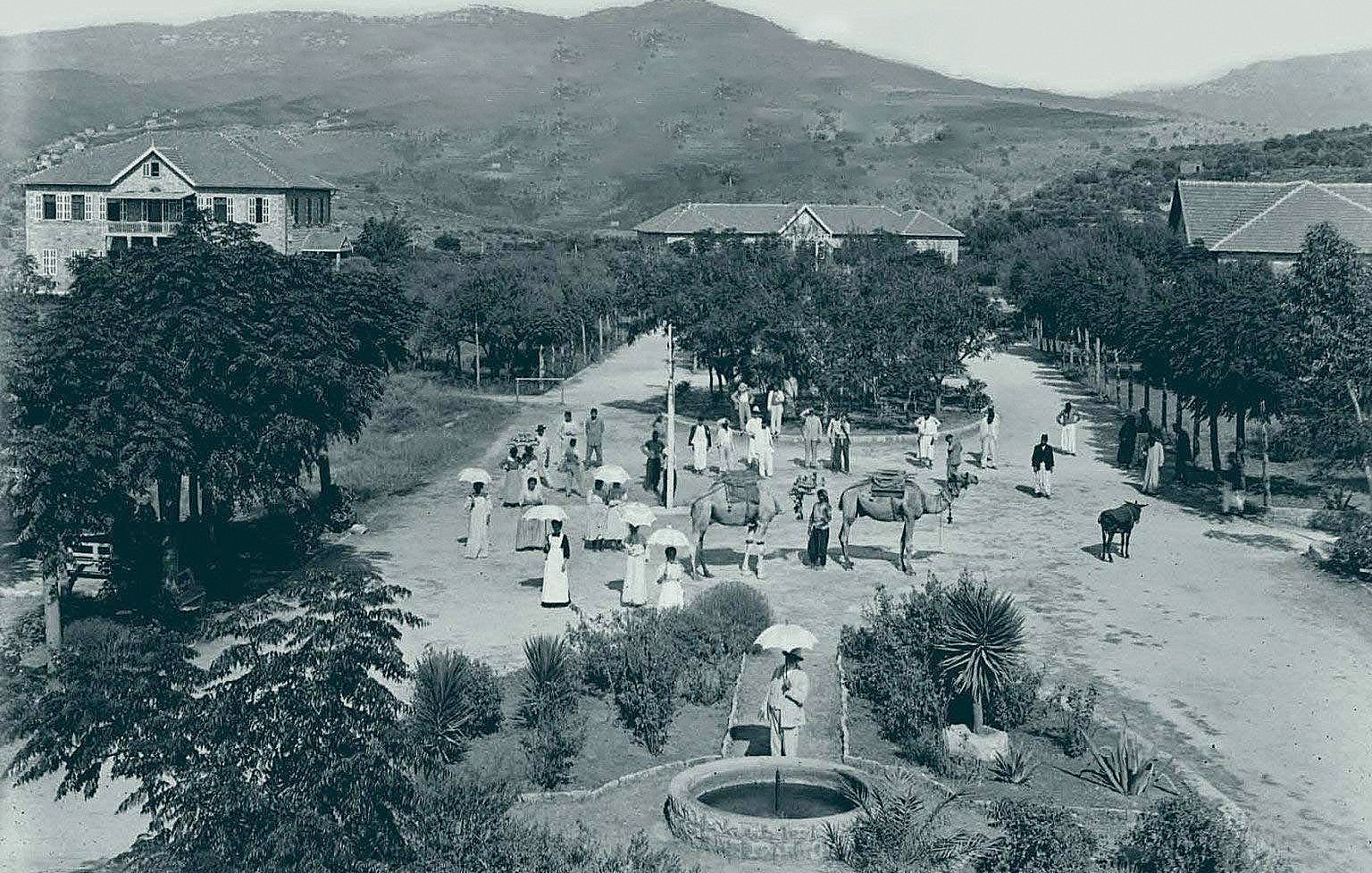 L'image semble représenter une scène rurale d'une époque passée. On peut voir un chemin central bordé d'arbres et de bâtiments à l'arrière-plan. Plusieurs personnes, vêtues de manière traditionnelle, se déplacent, certaines portant des parapluies. Des animaux comme des bœufs sont également présents, ajoutant à l'atmosphère agricole de la scène. Au premier plan, il y a un puits, probablement utilisé par les villageois. Les montagnes en arrière-plan complètent le paysage verdoyant. Cette image évoque une vie paisible et communautaire en harmonie avec la nature.