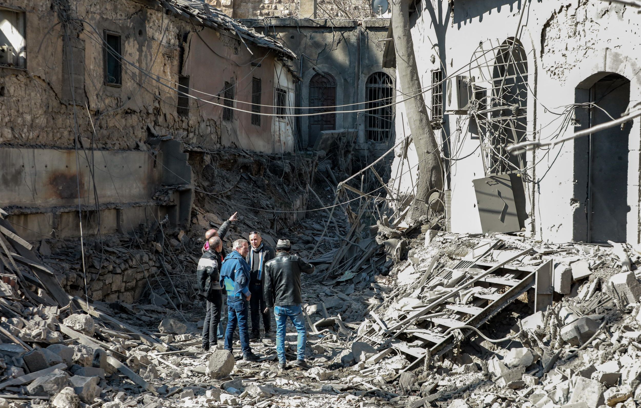 L'image montre un groupe de personnes se tenant au milieu d'un paysage de destruction. Les bâtiments autour d'eux sont en ruines, avec des murs effondrés et des débris éparpillés sur le sol. Les personnes semblent discuter ou examiner les lieux, tandis que des fils électriques sont visibles dans l'environnement. L'atmosphère est marquée par un sentiment de désolation et de perte.