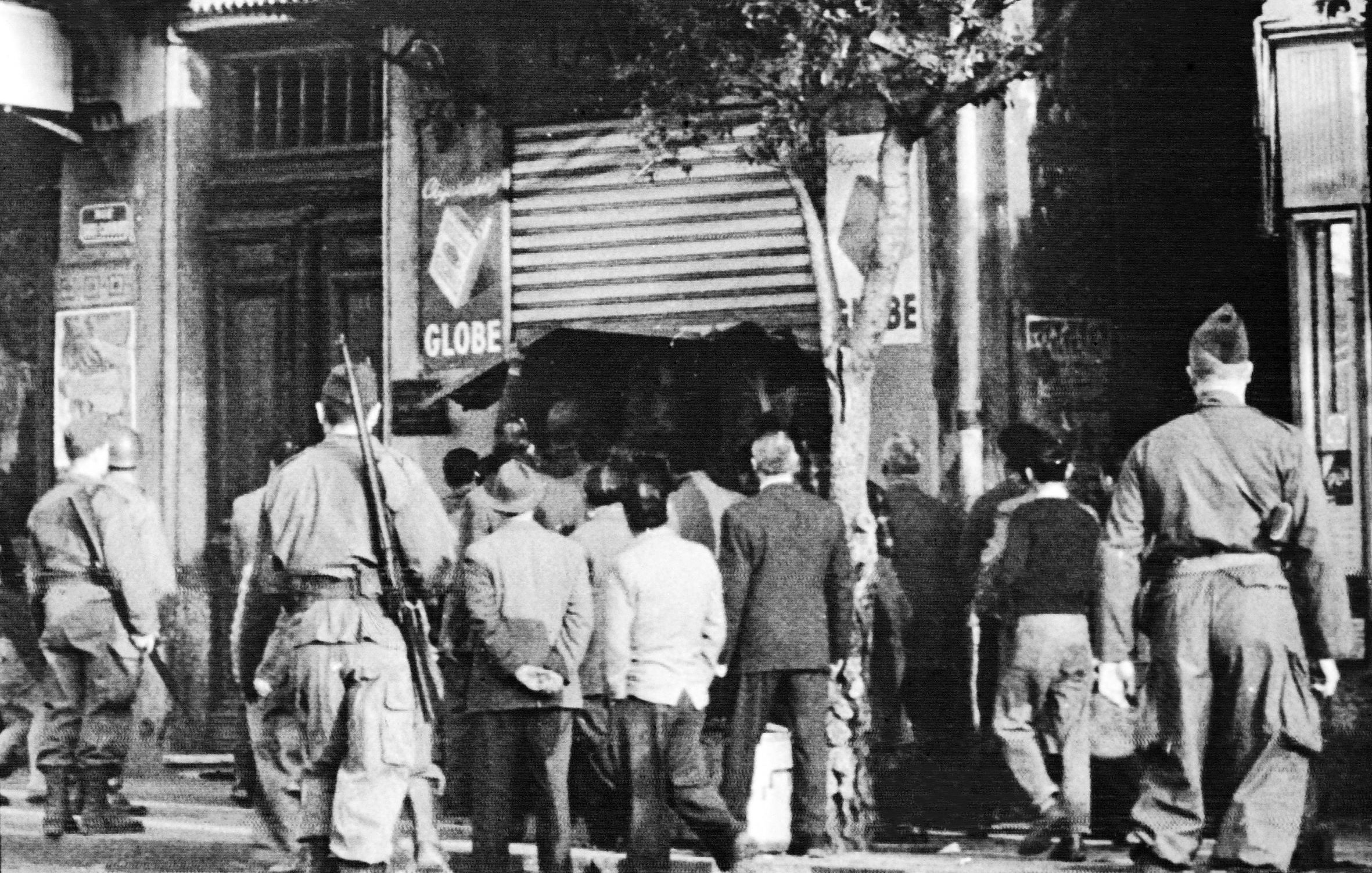 L'image montre une scène en noir et blanc où plusieurs soldats, en uniforme, se tiennent en arrière-plan. Devant eux, un groupe d'hommes en civil semble observer une situation à l'entrée d'un magasin ou d'un bâtiment, probablement une affaire en cours. La présence des soldats suggère un contexte de tension, peut-être lié à des événements historiques. Les détails de l'environnement, tels que les enseignes et l'architecture, évoquent une atmosphère urbaine, mais l'image elle-même semble figée dans le temps, ce qui renforce le caractère historique de la scène.