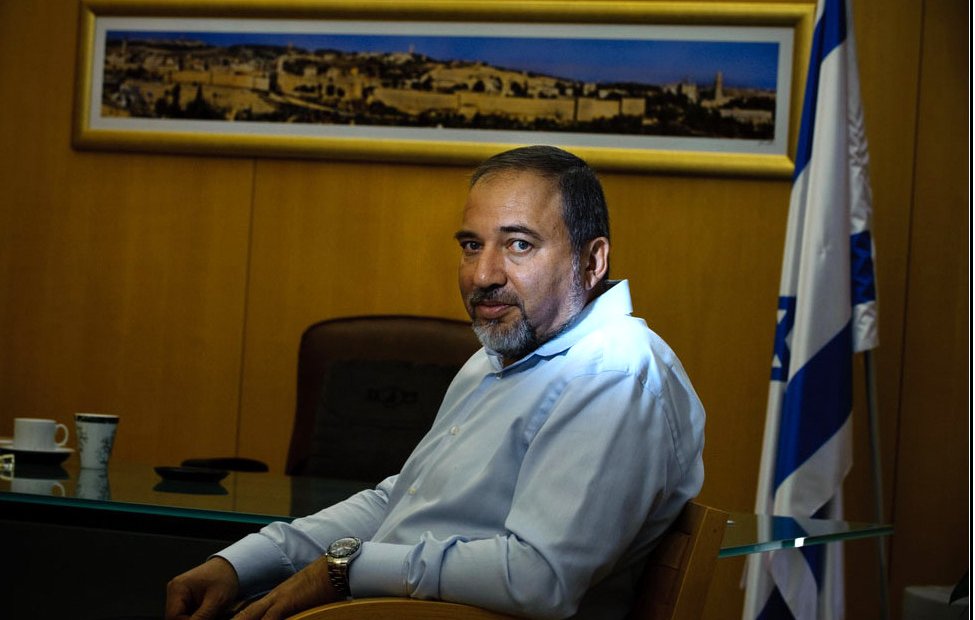 L'image montre un homme assis à un bureau. Il porte une chemise claire et a une expression sérieuse. En arrière-plan, on peut voir une grande photo d'une ville, probablement une vue de Jérusalem, et un drapeau, suggérant un contexte officiel ou politique. Le décor est sobre, avec une table en verre et une tasse sur le bureau.