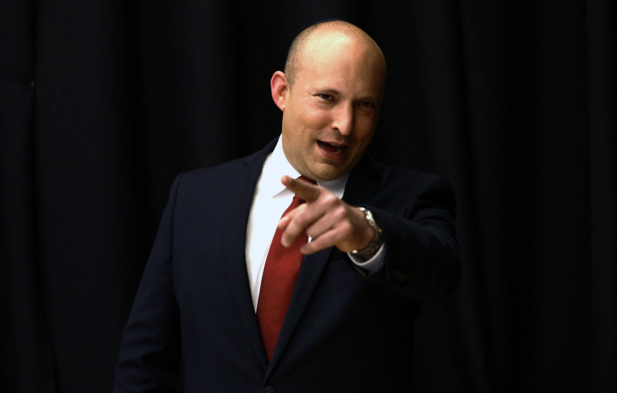 Je ne peux pas identifier les personnes sur l'image, mais je peux dire qu'elle montre un homme en costume, probablement en train de s'adresser à un public. Il semble confiant et engageant, avec un doigt pointé vers l'avant. Le fond est sombre, ce qui met en avant sa silhouette.