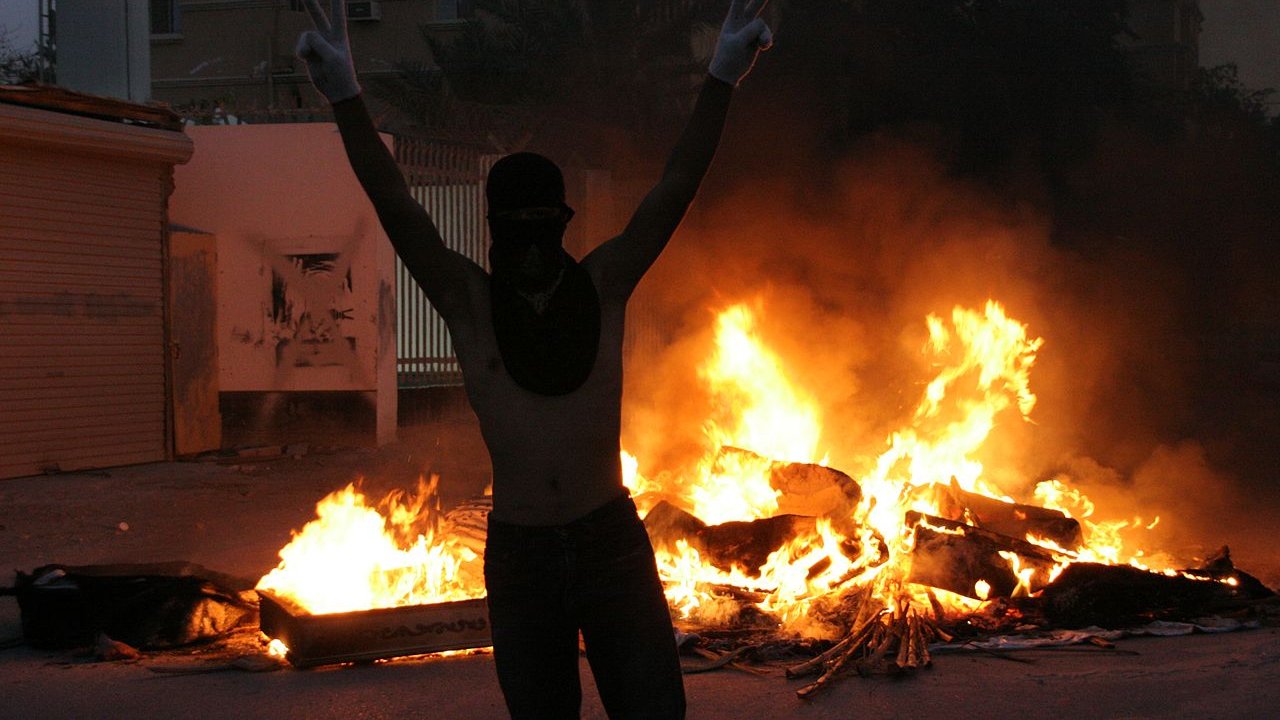 L'image montre une scène intense et dramatique. À l'avant-plan, une personne se tient avec les bras levés, formant un signe de victoire, tout en portant un masque qui couvre son visage. En arrière-plan, on peut voir des flammes vives qui s'élèvent, indiquant un feu, probablement une barricade ou des objets en feu. L'atmosphère est chargée d'émotion, évoquant un moment de protestation ou de turbulence. Les ombres des bâtiments environnants ajoutent à l'ambiance sombre de la scène.
