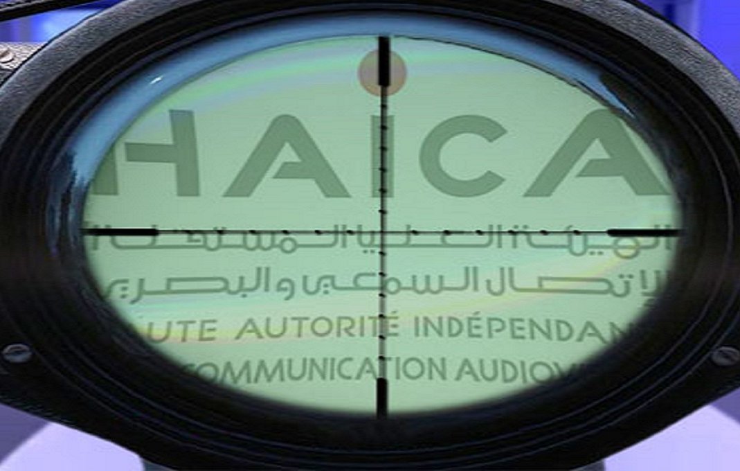 L'image montre un réticule de visée, typique d'une lunette de tir, qui se superpose à un fond avec le mot "HAICA" visible. Ce mot est associé à une autorité indépendante en matière de communication auditive, peut-être en rapport avec la régulation des médias. Le contraste entre l'optique et le texte suggère un thème de surveillance ou de contrôle. Le fond semble comporter divers éléments, peut-être liés à des événements ou des institutions médiatiques.