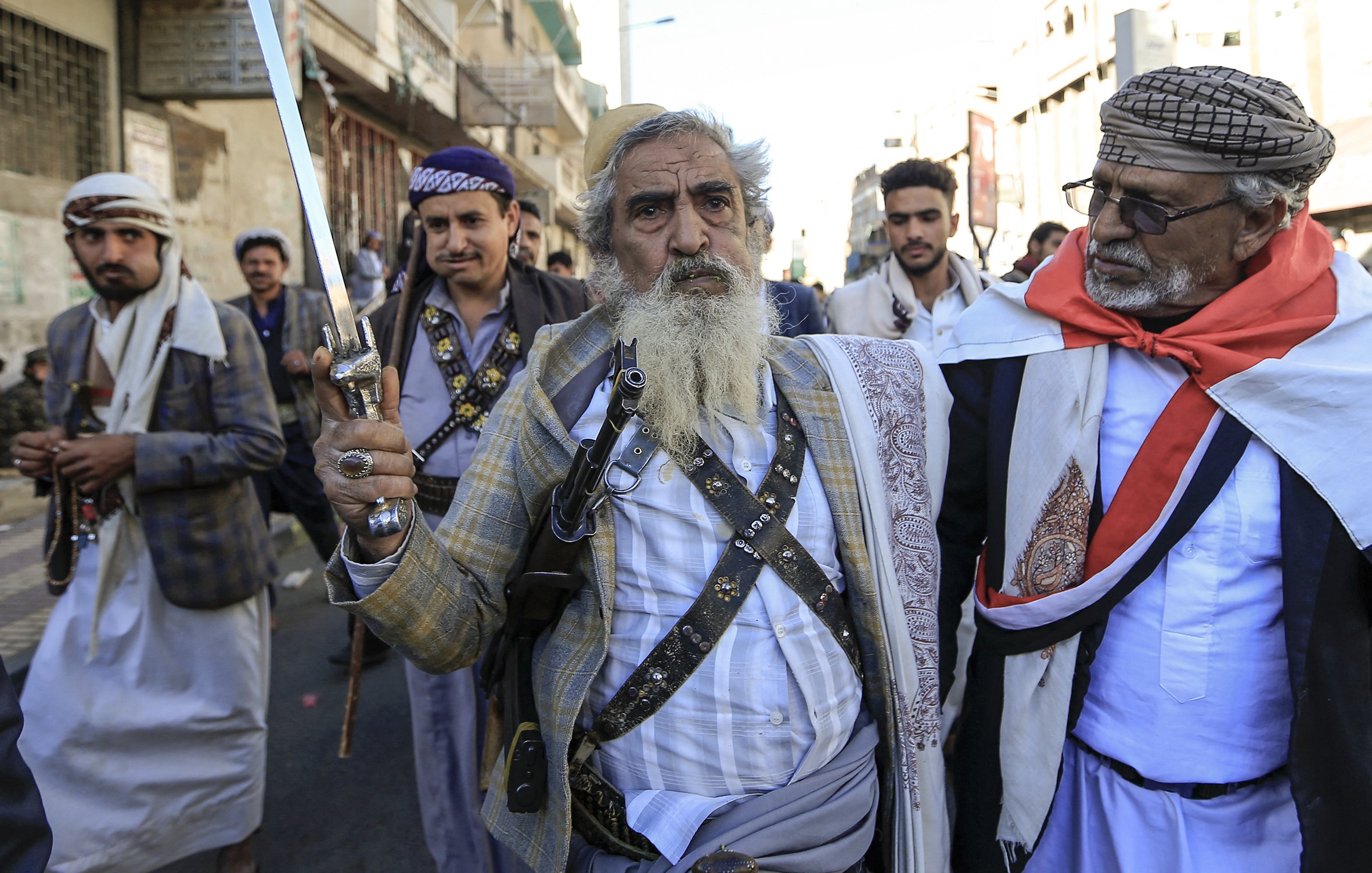 L'image montre un groupe d'hommes marchant dans une rue. L'un d'eux, au centre, est une personne âgée avec une longue barbe grise, tenant une épée. Il porte une tenue traditionnelle composée d'une chemise et d'une veste, ainsi qu'une ceinture. Les autres hommes dans le groupe portent aussi des vêtements traditionnels, et certains semblent porter des accessoires tels que des médaillons ou des fusils. L'ambiance de l'image suggère une manifestation ou une célébration culturelle. L'arrière-plan montre des bâtiments urbains typiques.