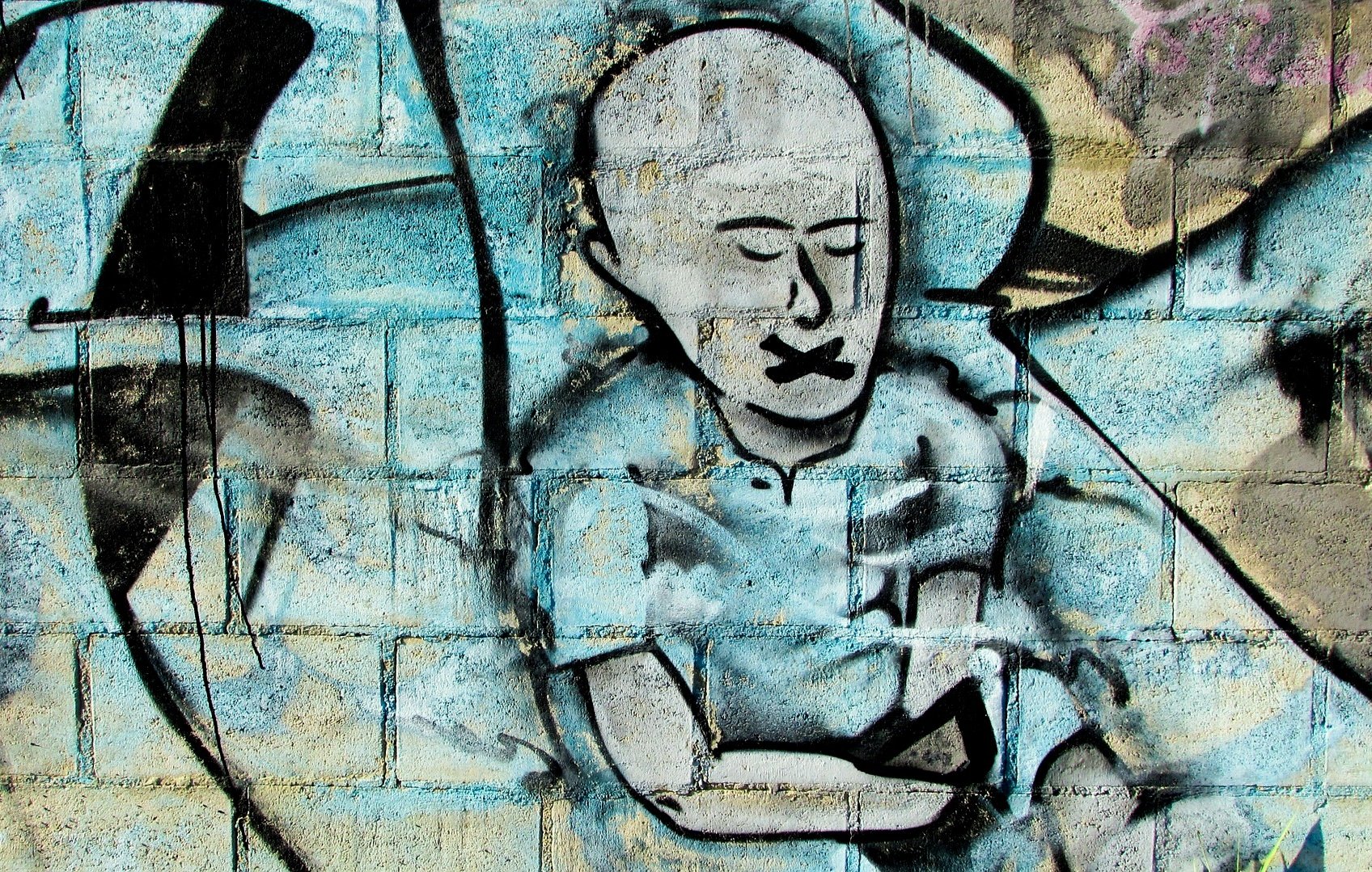 L'image présente un graffitti sur un mur. On y voit le dessin d'une figure humaine stylisée, sans cheveux, avec un visage exprimant le silence, probablement pour symboliser l'oppression ou le manque de voix. Ses lèvres sont représentées comme étant scellées. Le fond est composé de couleurs bleu et gris, avec des formes abstraites qui ajoutent de la texture à l'œuvre. L'ensemble dégage une ambiance à la fois poignante et réfléchie.