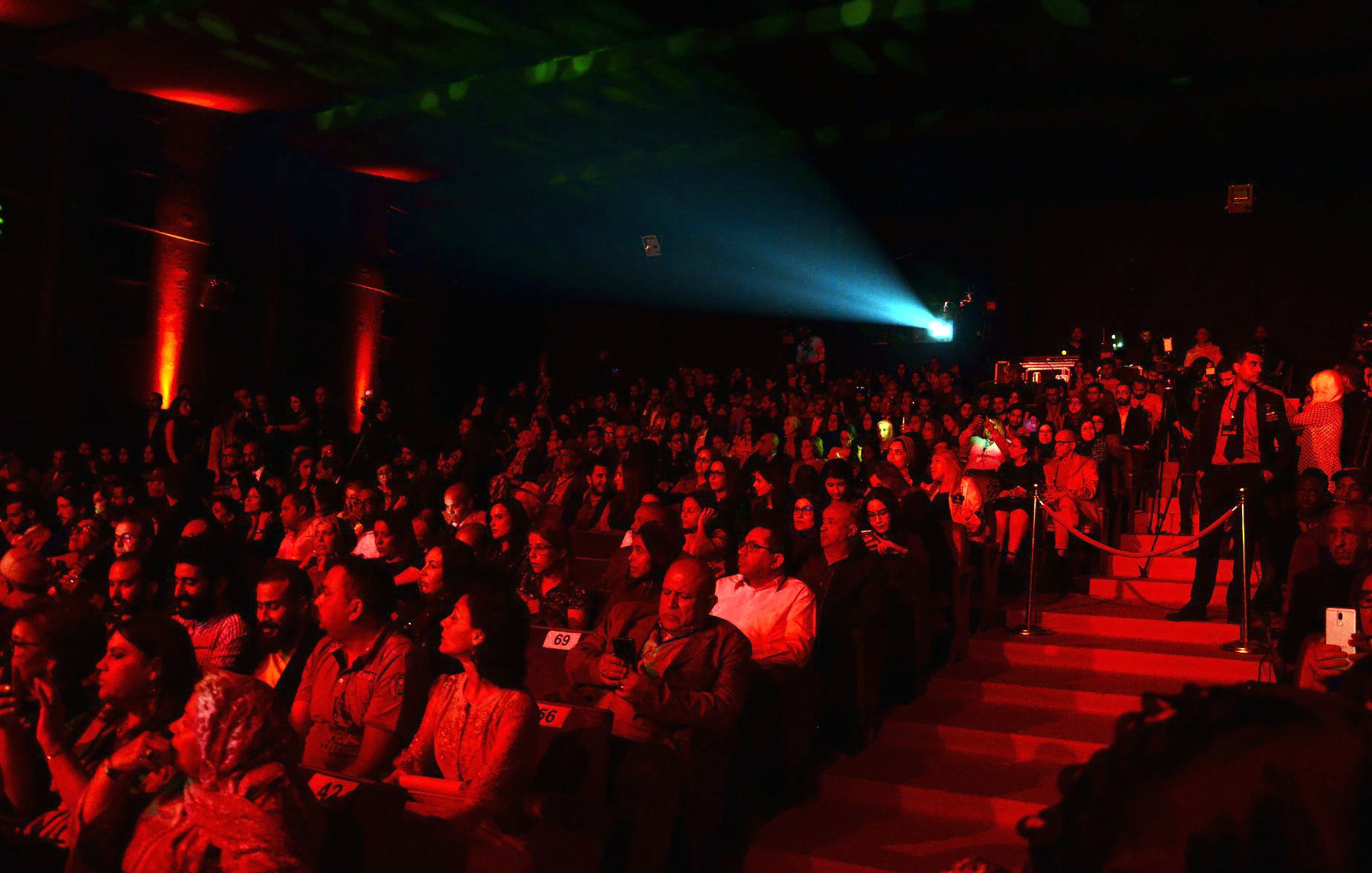 L'image montre un auditorium rempli de spectateurs. La salle est faiblement éclairée avec des lumières rouges qui créent une ambiance chaleureuse et dynamique. Au centre, un projecteur émet un faisceau lumineux, ajoutant à l'atmosphère du moment. Les spectateurs, composés d'hommes et de femmes, semblent attentifs et engagés, certains tenant des téléphones. L'escaliers sur le côté de la scène permet l'accès à d'autres niveaux. L'ensemble de la scène évoque un événement artistique ou culturel.