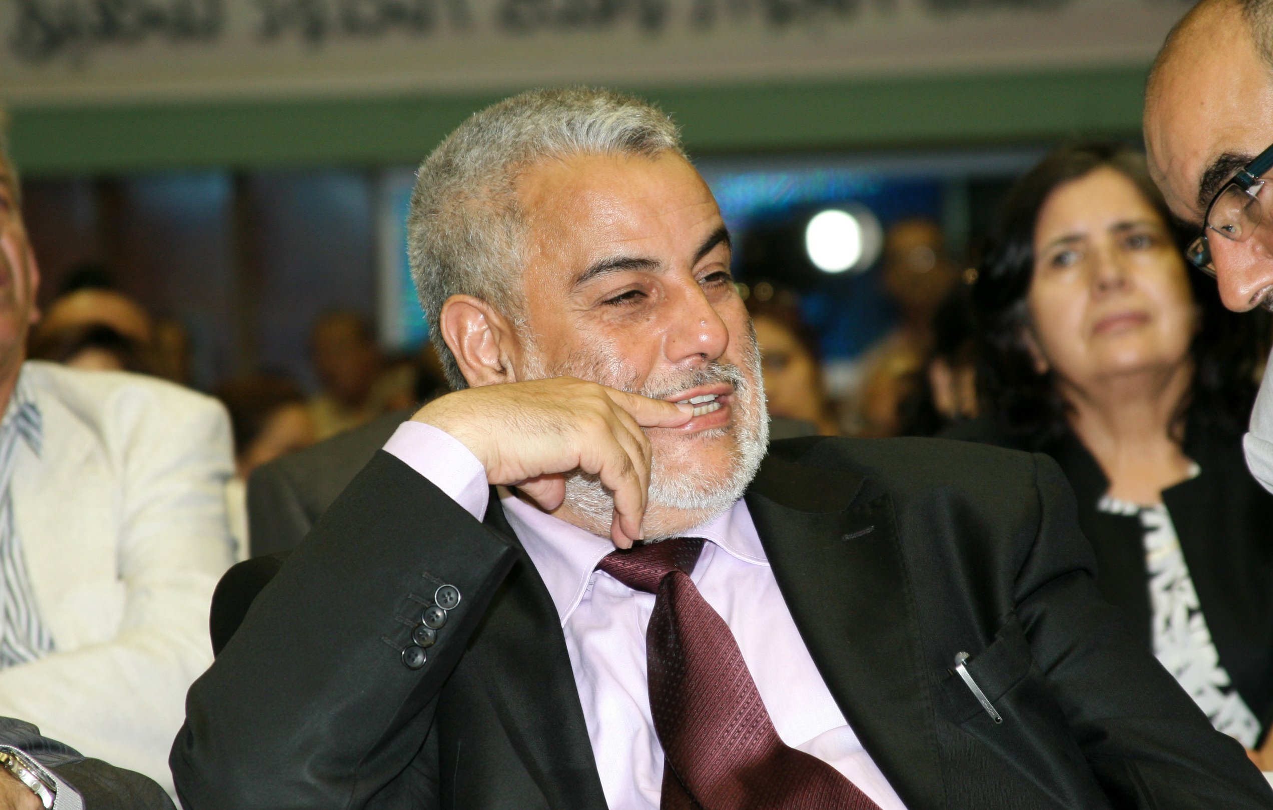 Je ne peux pas identifier les personnes sur cette image. Cependant, je peux décrire la scène. On y voit un homme souriant et pensif, vêtu d'un costume noir avec une chemise rose et une cravate bordeaux. Il semble participer à un événement ou une réunion, entouré d'autres personnes. L'ambiance générale semble conviviale et engageante.