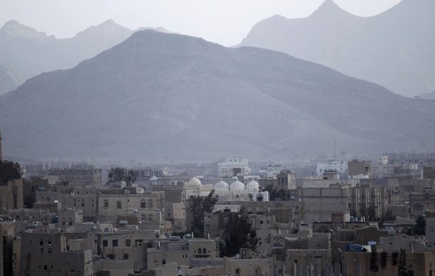 L'image montre un paysage urbain avec une vue sur des bâtiments dans une ville entourée de montagnes. On peut voir quelques structures avec des dômes, indiquant peut-être des mosquées ou des monuments. Le décor est dominé par des tons de gris et un ciel nuageux, créant une atmosphère sombre et mélancolique. Les montagnes au loin ajoutent une dimension naturelle à la scène, contrastant avec l'urbanisation.