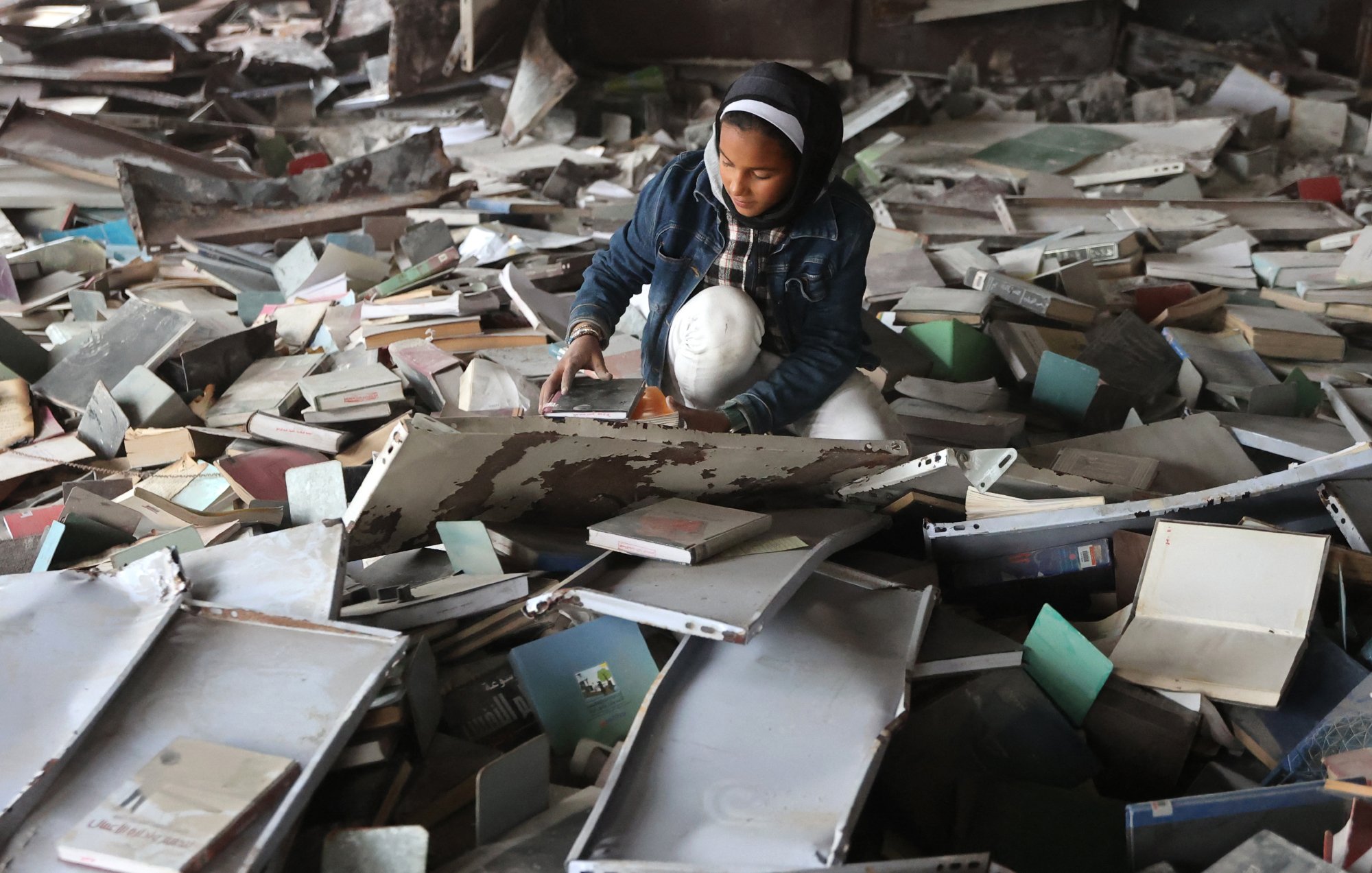 L'image montre une jeune fille assise au milieu d'un tas de livres et de débris. Elle semble explorer et manipuler les objets autour d'elle dans un environnement en désordre.