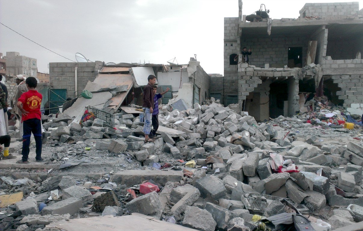 L'image montre des bâtiments gravement endommagés, avec des débris éparpillés sur le sol. On peut voir des enfants et des adultes présents parmi les ruines, cherchant peut-être des effets personnels ou évaluant les dégâts. L'environnement semble chaotique et témoigne d'une destruction significative, probablement suite à un conflit ou à une catastrophe. Le ciel est nuageux, ce qui ajoute à l'atmosphère sombre de la scène.