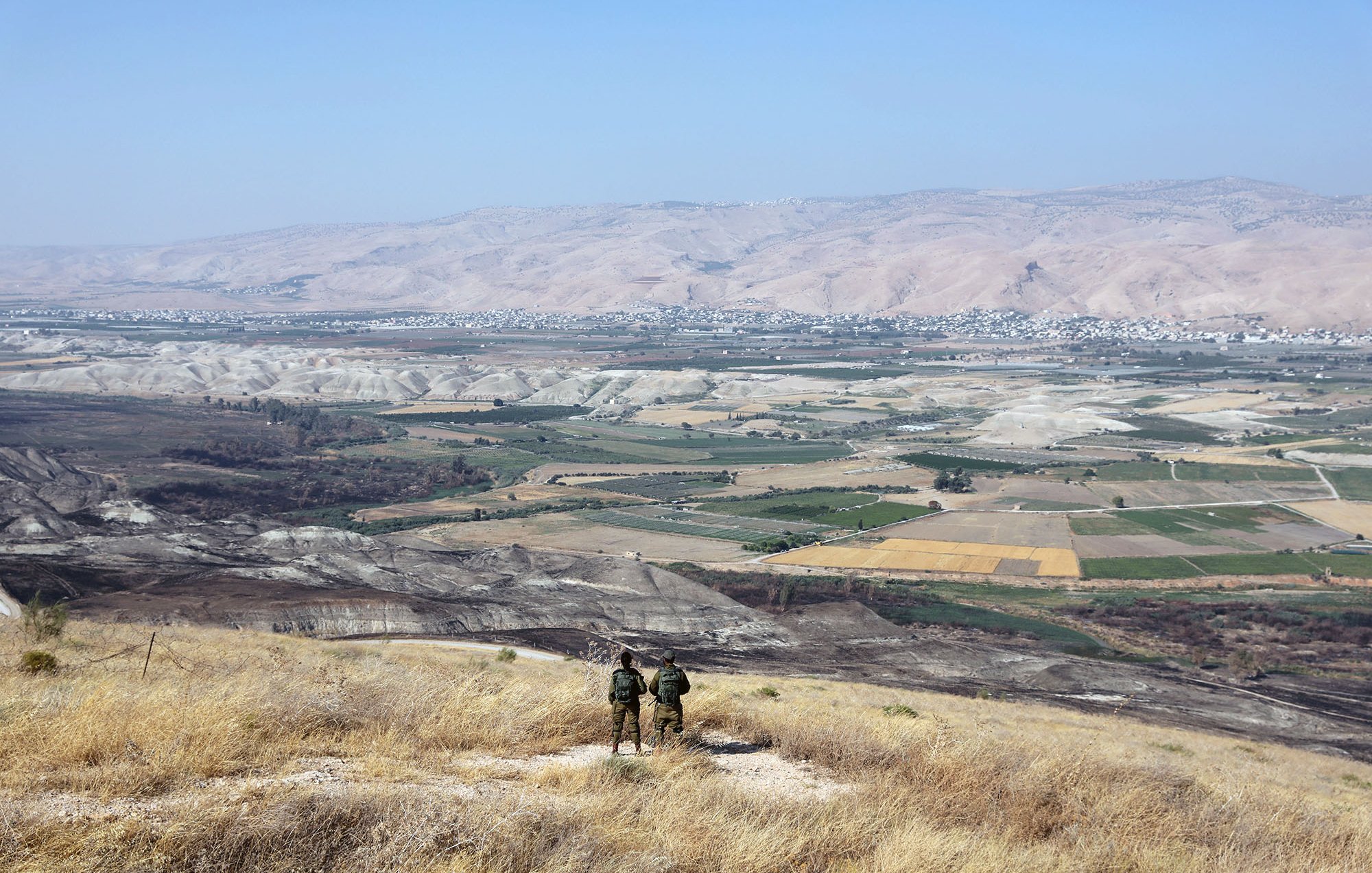 L'image montre un paysage montagneux et rural. Au premier plan, deux personnes se tiennent de dos, probablement des soldats ou des randonneurs, observant la vallée en contrebas. La vallée est composée de champs et de terrains cultivés, avec des couleurs variées allant du vert au jaune. À l'arrière-plan, des collines et des montagnes se profilent sous un ciel bleu clair. L'ensemble dégage une impression de tranquillité et de vaste espace naturel.