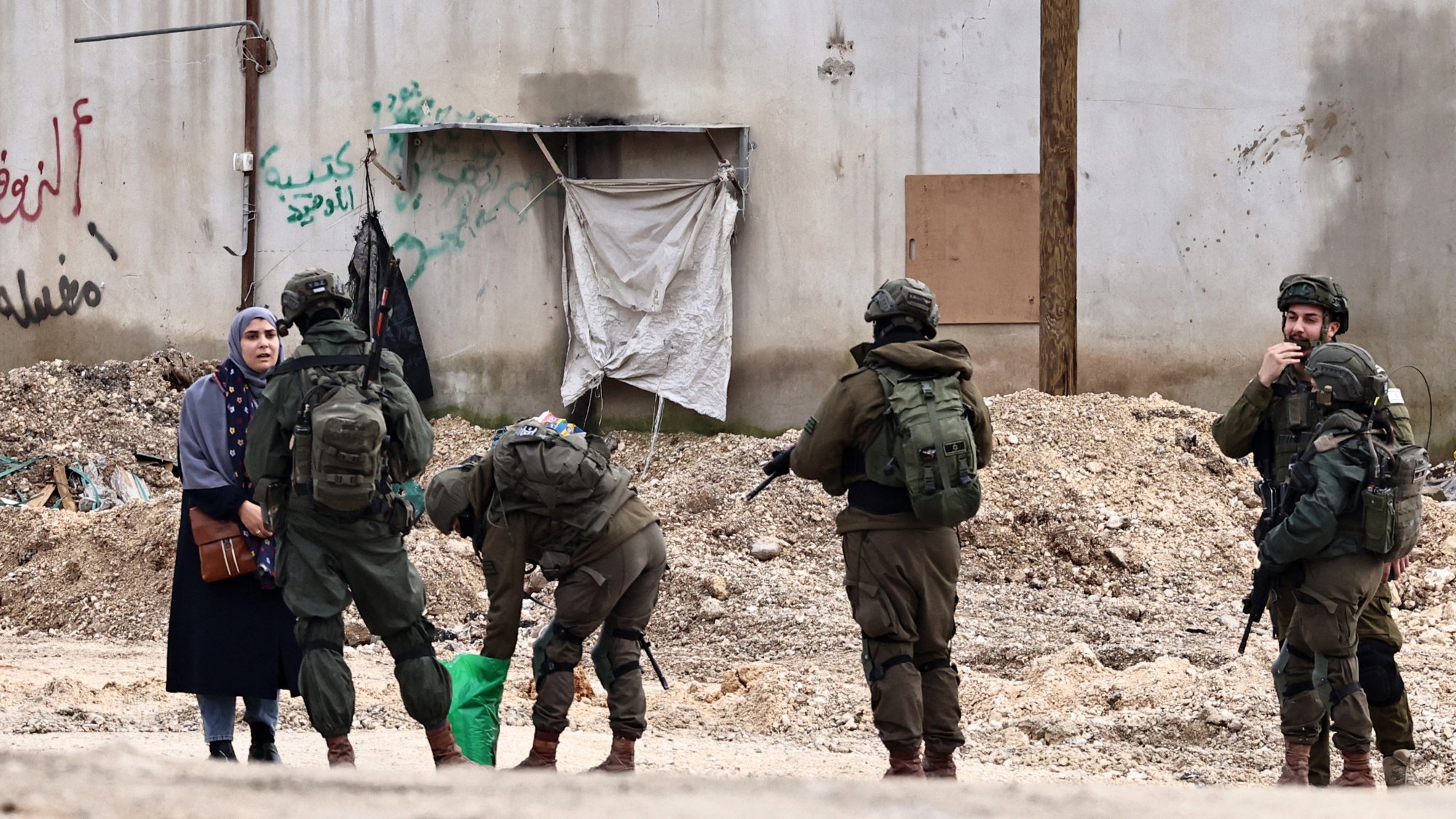 L'image montre une femme vêtue d'une robe sombre, se tenant à côté d'une rue. Devant elle, plusieurs soldats en uniforme sont regroupés, certains sont en train de fouiller un sac, tandis que d'autres observent la femme. En arrière-plan, on peut voir un mur avec des graffitis et des débris sur le sol.