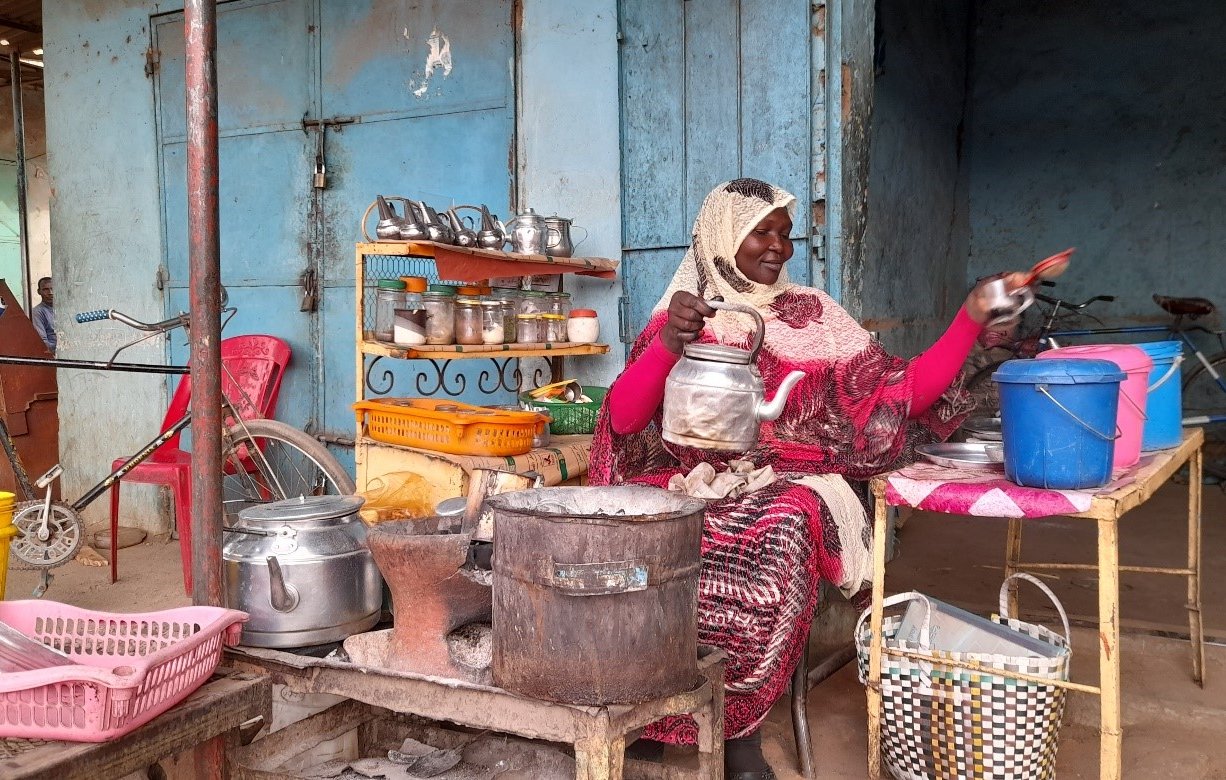 L'image montre une femme assise devant un petit stand ou un kiosque. Elle porte un vêtement traditionnel, probablement coloré, et a un foulard sur la tête. Autour d'elle, on peut voir divers ustensiles de cuisine et des récipients, comme des casseroles et des seaux en plastique de différentes couleurs. Sa table est en métal et semble utilisée pour préparer ou vendre de la nourriture. En arrière-plan, on aperçoit des murs peints et d'autres objets, créant une atmosphère vivante et locale.