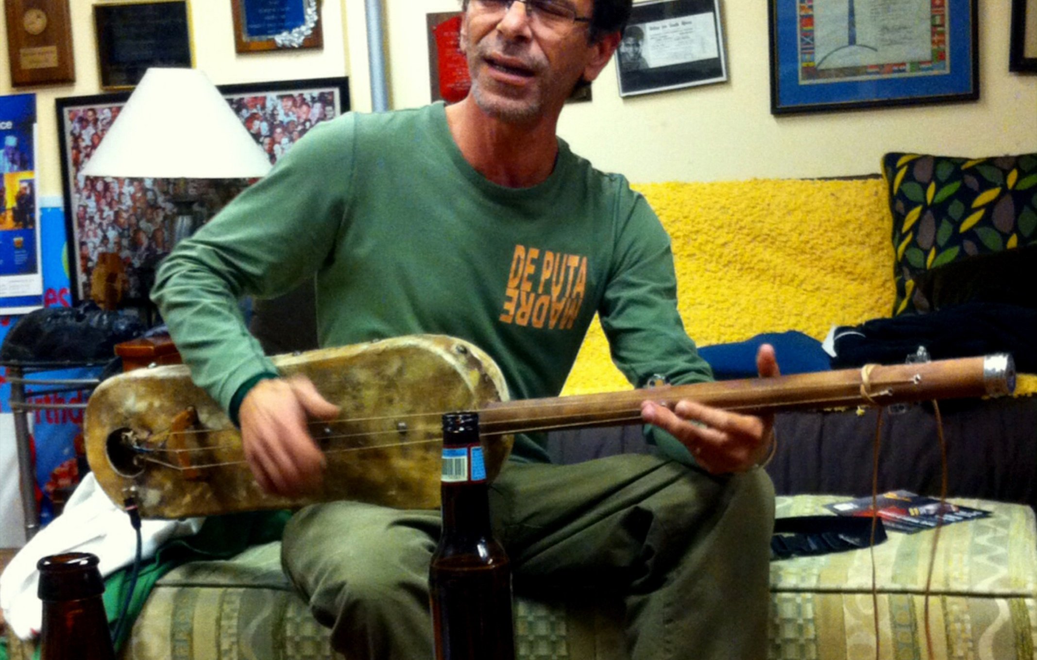 L'image montre un homme assis dans un intérieur chaleureux, probablement un salon. Il joue d'un instrument de musique, semblable à une guitare, tout en chantant. Il porte un t-shirt avec un message audacieux et des pantalons clairs. À côté de lui, sur une table basse, on aperçoit quelques bouteilles de bière. Les murs autour sont décorés de plaques et de diplômes, et un canapé coloré est visible en arrière-plan. L'ambiance semble conviviale et détendue.