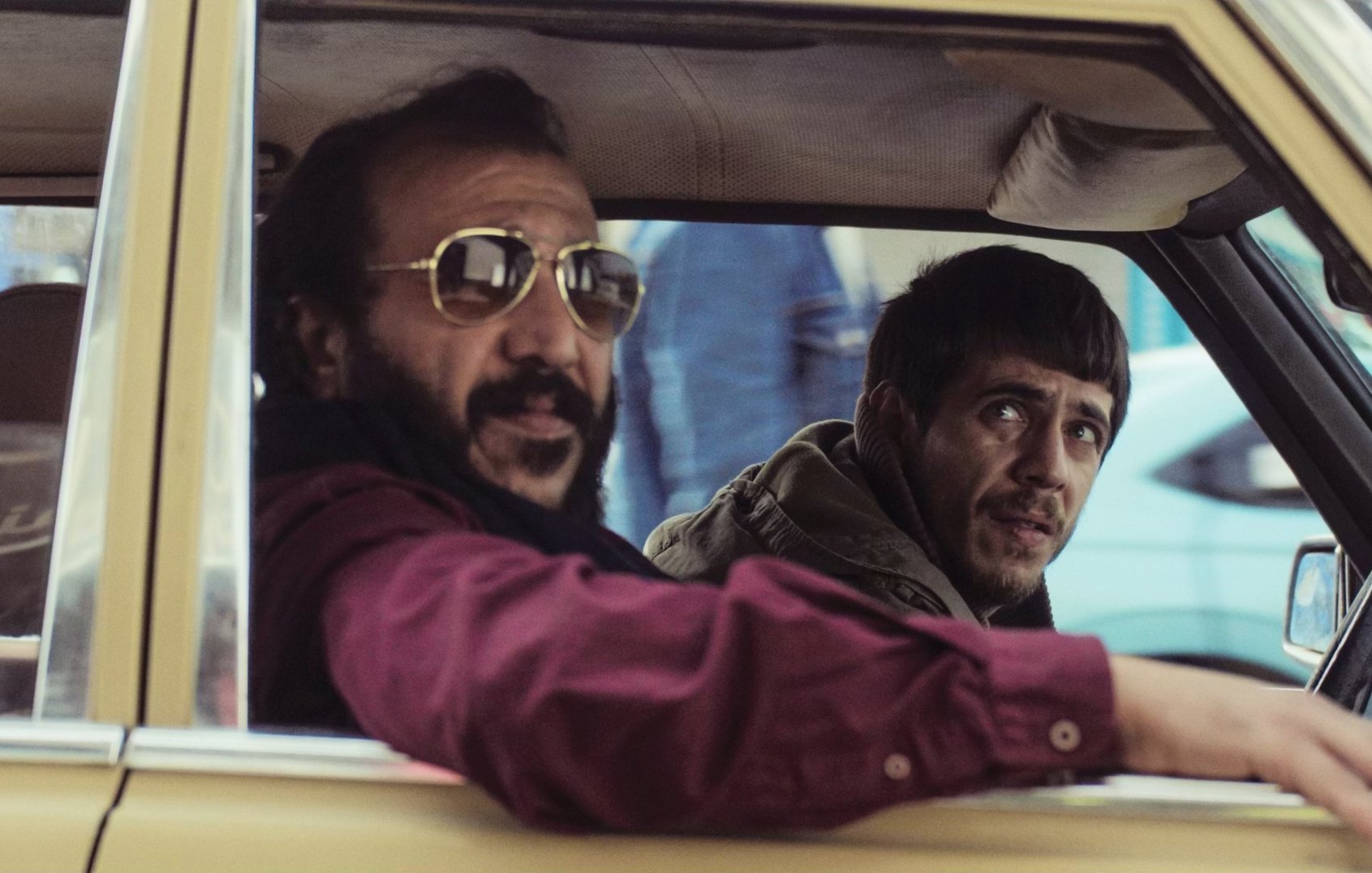 Deux hommes dans une voiture, l'un avec des lunettes de soleil, l'autre semble inquiet.