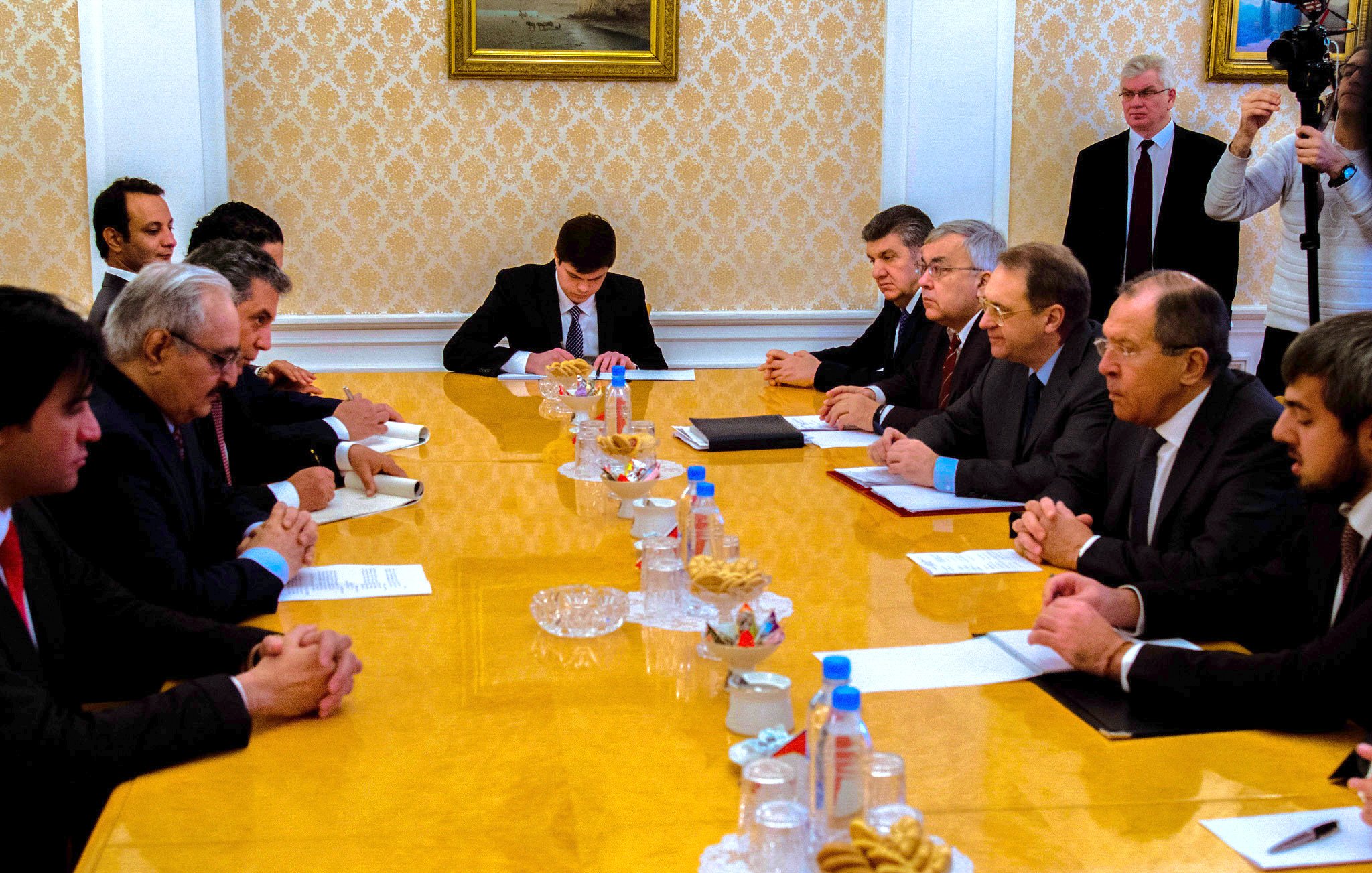 L'image présente une réunion autour d'une grande table ovale en bois. Plusieurs hommes sont assis des deux côtés, en tenue formelle, semblant être en discussion ou en train de négocier. Au centre de la table, il y a des documents et des canapés, ainsi qu'une assiette de collations. En arrière-plan, on peut voir des personnes observant la réunion, ce qui indique que c'est un moment important. L'atmosphère semble sérieuse et concentrée.