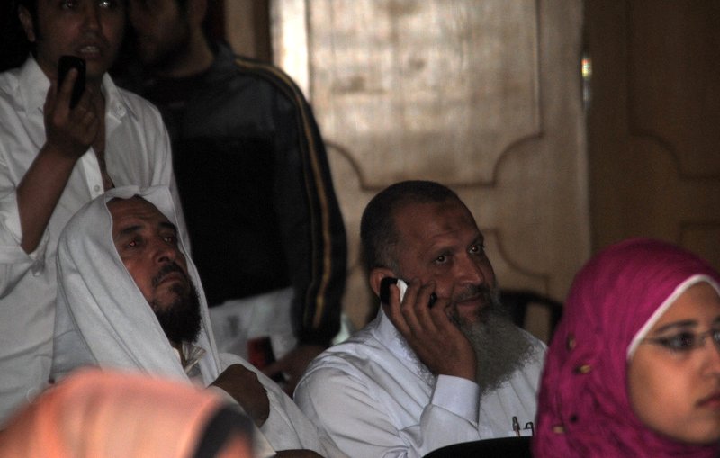 L'image montre des hommes et des femmes dans un environnement qui semble être un rassemblement ou une conférence. Deux hommes au premier plan portent des vêtements traditionnels, l'un d'eux est exprimé en train de passer un coup de téléphone. L'arrière-plan montre d'autres personnes, certaines concentrées sur l'événement, et des couleurs variées dans les vêtements, y compris une femme portant un hijab rose. L'atmosphère semble sérieuse et attentive.