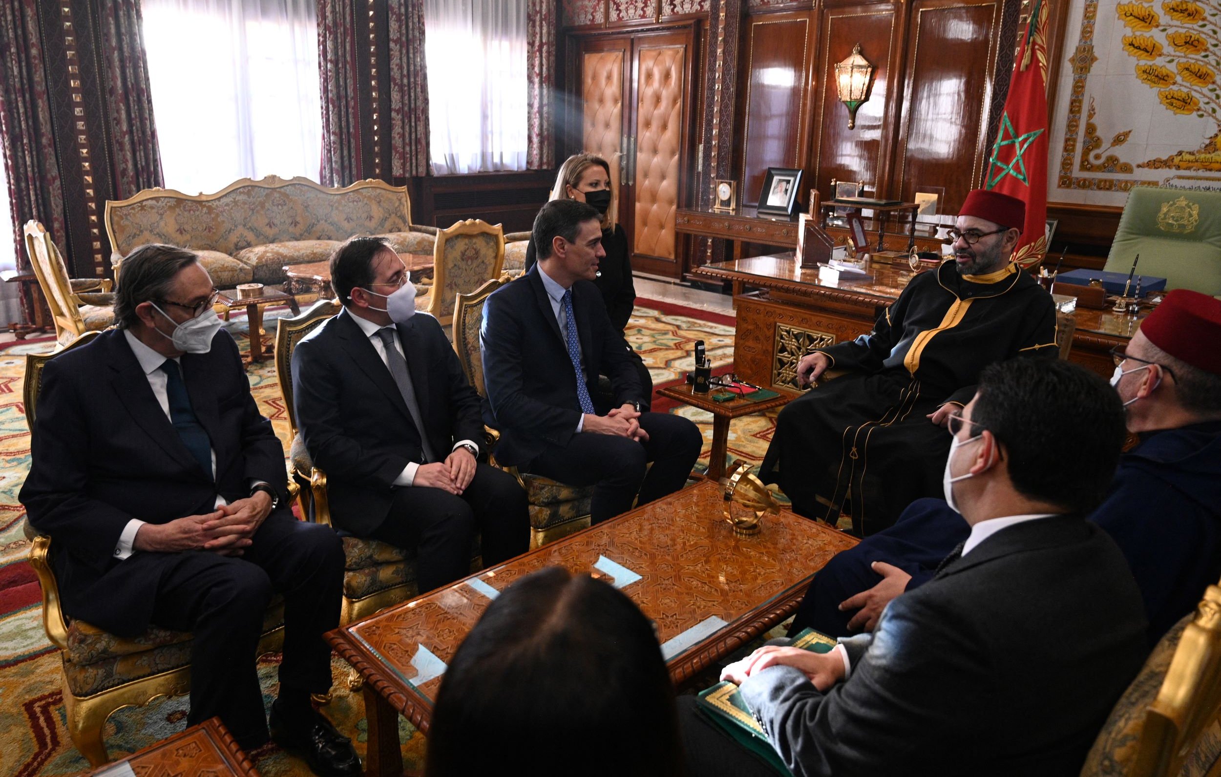 L'image montre une réunion dans un cadre officiel, probablement un bureau ou une salle de réunion importante. Plusieurs personnes en costumes sont assises autour d'une table, tandis qu'une autre personne, vêtue d'une tenue traditionnelle, est assise en face d'eux. Les participants portent des masques, ce qui indique que la réunion pourrait avoir lieu en période de pandémie. L'environnement est richement décoré, avec des éléments de design marocain et un mobilier en bois orné.