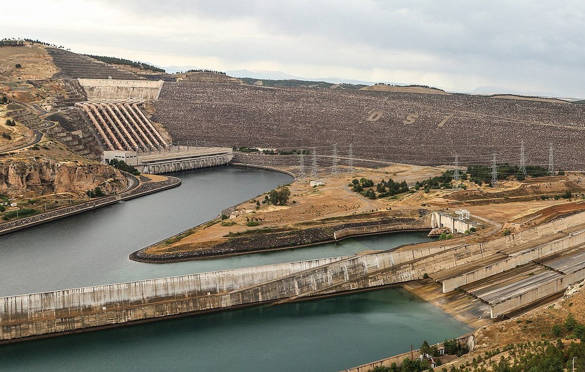 L'image montre une vue panoramique d'un complexe de barrages ou d'une centrale hydroélectrique. On peut voir un grand réservoir d'eau entouré par des collines sur lesquelles se trouvent des structures en béton. Des canaux ou des tunnels d'acheminement de l'eau sont visibles, ainsi que des installations de contrôle des niveaux d'eau. Le paysage est principalement aride, avec quelques zones verdoyantes et une atmosphère dramatique due à un ciel nuageux. Les infrastructures semblent bien entretenues et sont probablement liées à la production d'énergie.