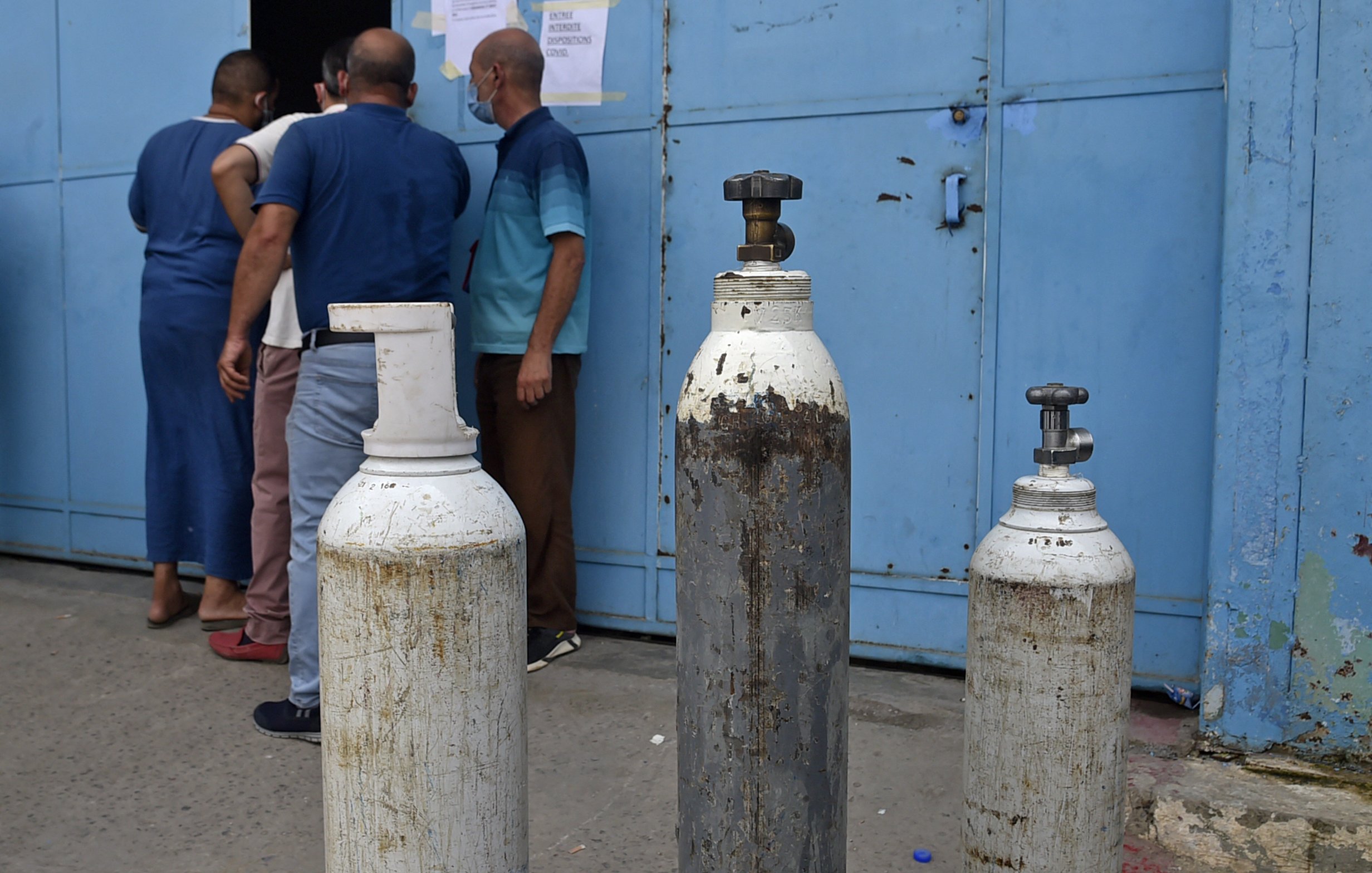 L'image montre un groupe de personnes devant une porte bleue. Sur le devant, il y a plusieurs bouteilles de gaz, dont certaines sont blanches et d'autres en métal. Les bouteilles semblent usées et peuvent être utilisées pour stocker des gaz, comme l'oxygène. Les personnes à l'arrière semblent attendre ou discuter, tandis que l'environnement semble être un lieu urbain.