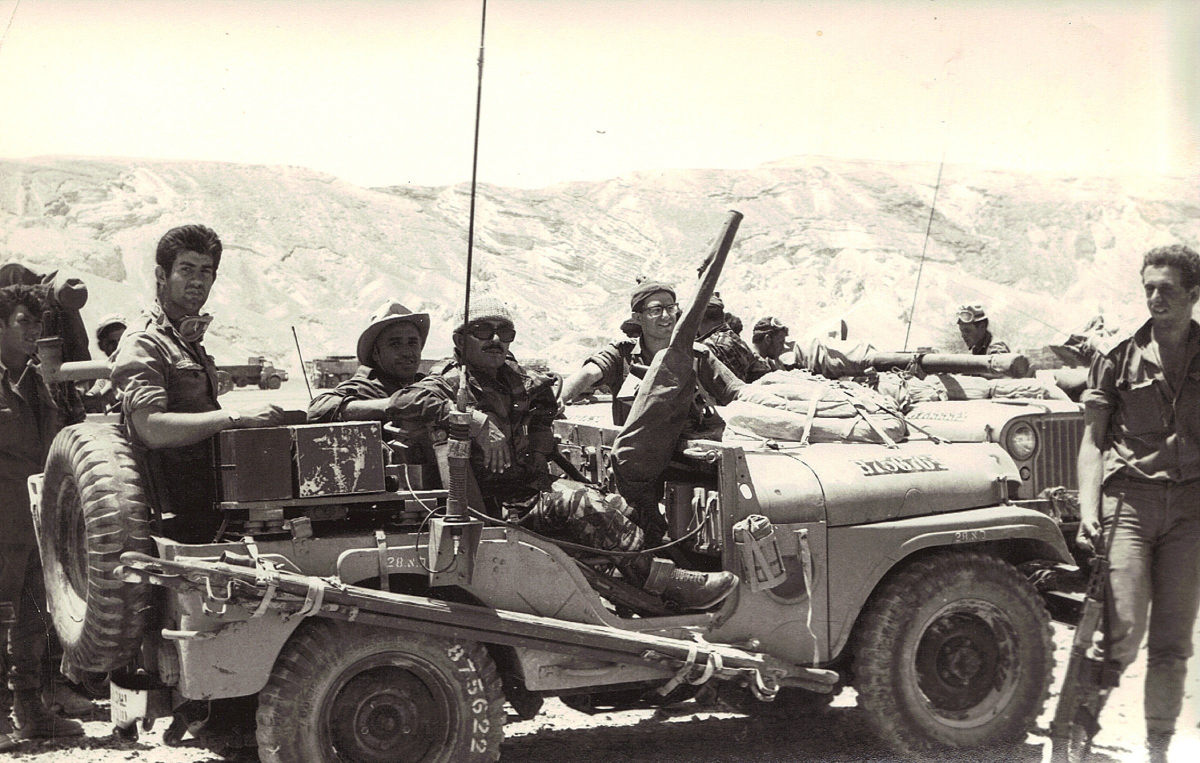 L'image montre un groupe de soldats réunis autour d'un véhicule militaire, probablement une jeep. Ils semblent engagés dans une mission, avec des équipements militaires visibles sur le véhicule. Le paysage en arrière-plan est aride, avec des collines ou des montagnes. Les soldats prennent la pose, certains souriant, et semblent confiants dans leur environnement. L'atmosphère dégage un sentiment de camaraderie et de détermination.