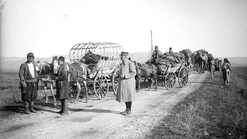 L'image montre une scène historique sur un chemin. On y voit plusieurs chariots tirés par des chevaux, ainsi que des personnes qui se déplacent. Certaines personnes sont debout près des chariots, tandis que d'autres semblent s'affairer autour des animaux. L'environnement est rural, avec des champs en arrière-plan et une atmosphère qui évoque des temps passés, probablement sur un chemin de terre. Les vêtements des personnes suggèrent une époque ancienne.