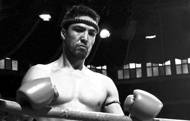 L'image montre un boxeur debout dans un ring. Il est torse nu et porte un bandeau autour de la tête. Ses poings sont protégés par des gants de boxe. Son expression semble concentrée et déterminée. Le style de l'image est en noir et blanc, ce qui lui donne une ambiance nostalgique et intemporelle. Le ring et l'arrière-plan suggèrent un environnement de compétition.