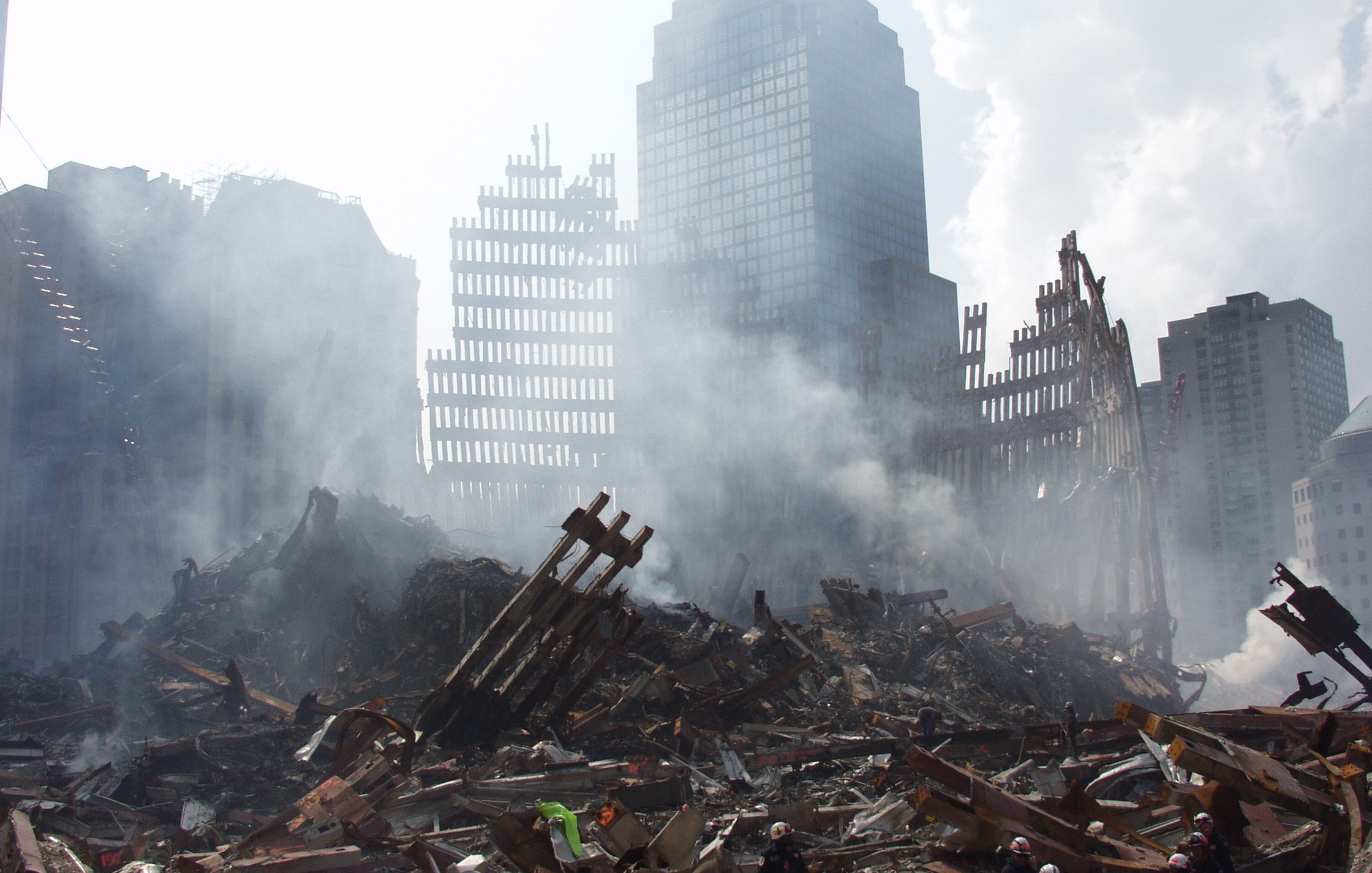 L'image montre un paysage urbain dévasté, avec des débris de bâtiments effondrés et des structures en ruines. De la fumée s'élève dans l'air, créant une atmosphère sombre et chaotique. On peut voir des restes de structures métalliques, avec des morceaux de béton éparpillés sur le sol. L'arrière-plan présente des gratte-ciel partiellement visibles, suggérant une zone qui a subi des dommages significatifs. La scène évoque un sentiment de destruction et de désolation.
