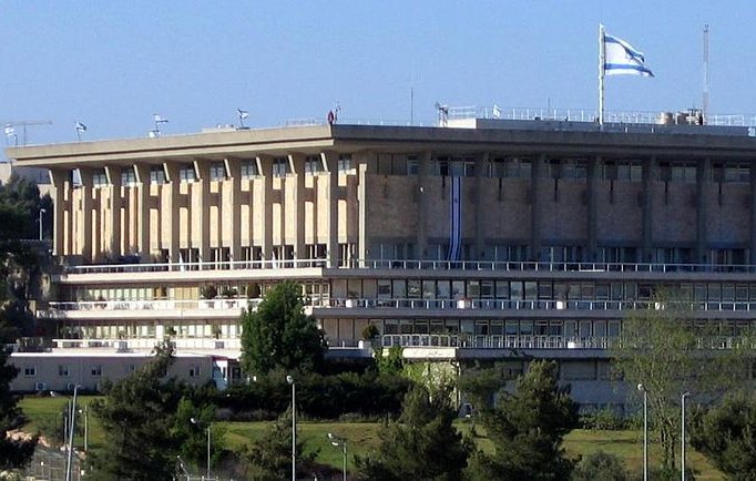 L'image montre un bâtiment moderne avec une architecture distincte, probablement situé dans un environnement urbain. Le bâtiment a plusieurs niveaux, avec des fenêtres et des balcons visibles. On peut également apercevoir des drapeaux, y compris celui d'Israël, flottant en haut du bâtiment. Autour, il y a des arbres et de la verdure, ce qui donne une impression de nature intégrée dans un cadre urbain. Le ciel est bleu et dégagé, ajoutant à la luminosité de la scène.