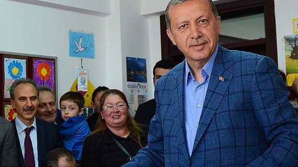 L'image montre un homme d'âge moyen portant un costume bleu. Il est debout, tenant un bulletin dans sa main, et semble être dans une salle de vote ou un endroit similaire. Il y a plusieurs personnes en arrière-plan, dont une femme avec des lunettes, un enfant et d'autres adultes, qui regardent l'homme avec intérêt. L'environnement est chaleureux et coloré, avec des affiches accrochées aux murs.