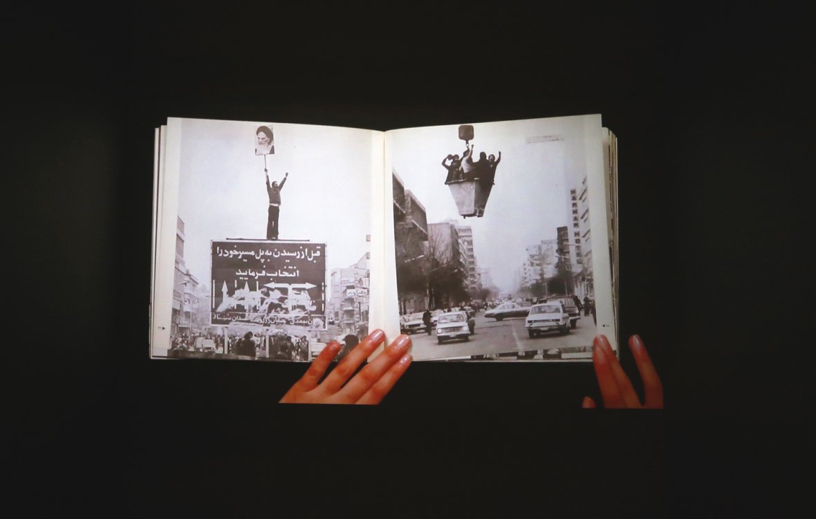 L'image montre un livre ouvert, tenu par des mains, avec des pages affichant des photos en noir et blanc. L'une des pages présente une grande pancarte ou affiche, tandis que l'autre illustre une rue avec des voitures. L'ensemble évoque des thèmes de mouvement, de protestation ou d'événements historiques, en jouant sur le contraste entre les images et le texte visibles. L'arrière-plan est sombre, ce qui met en valeur le livre et les illustrations qu'il contient.