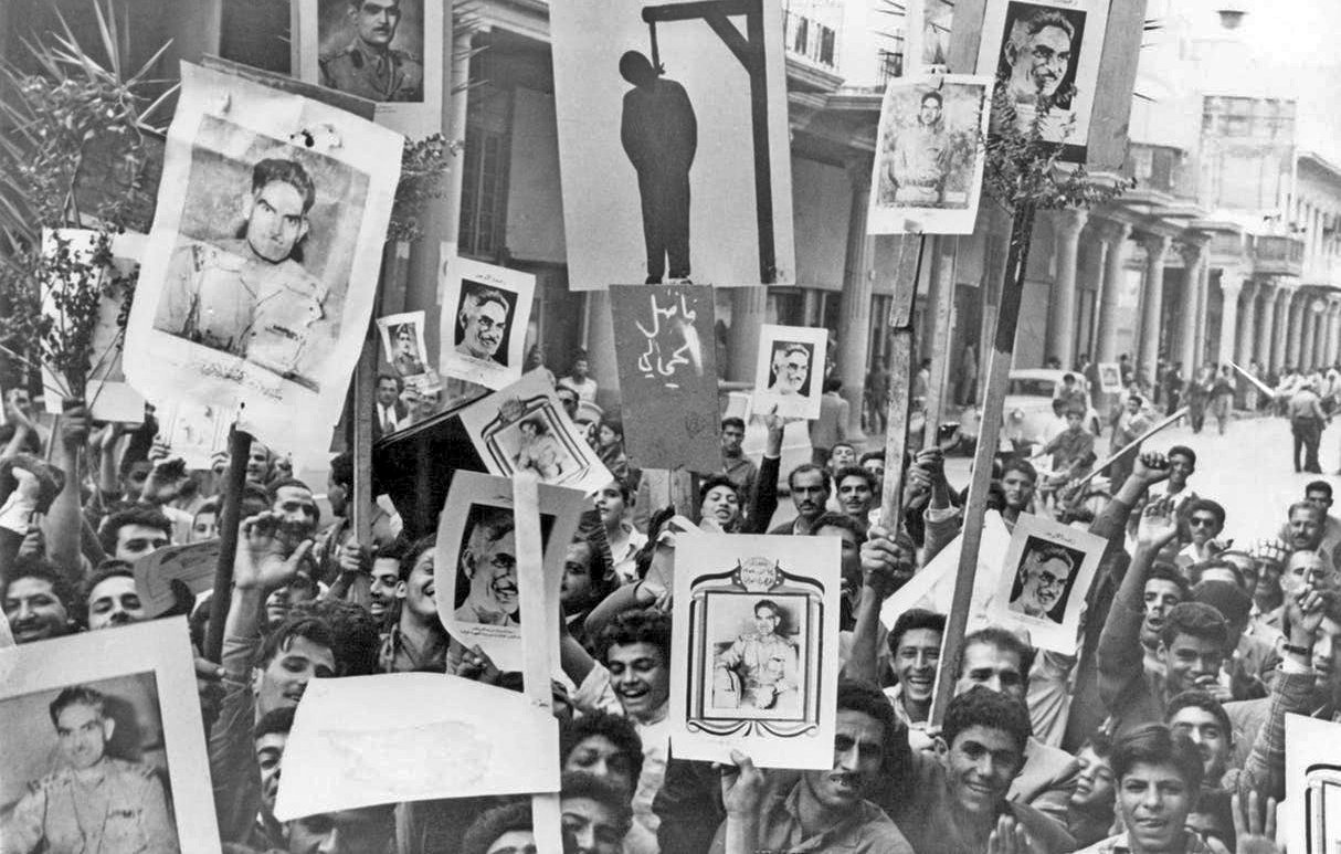 L'image montre une foule de personnes rassemblées dans une rue, tenant des pancartes et des portraits en protestation. On peut voir un dessin d'une potence au centre de l'affichage, suggérant un contexte de tension ou de révolte. Les visages des manifestants expriment diverses émotions, et l'environnement semble animé. Des palmiers sont visibles en arrière-plan, ajoutant une touche de verdure à la scène.