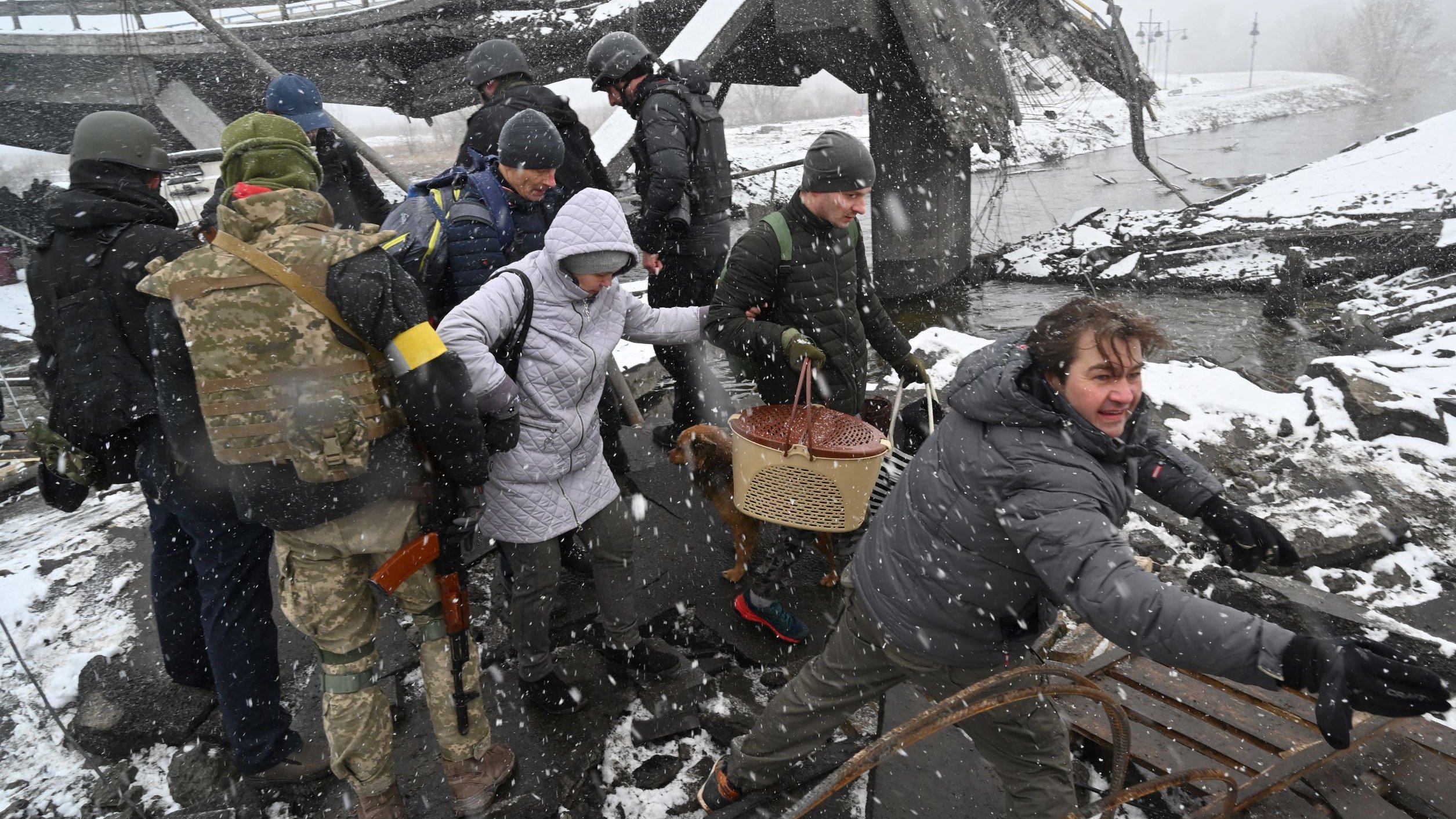 L'image montre un groupe de personnes en train d'évoluer dans un environnement de débris, probablement après un sinistre. Des soldats en uniforme sont présents, ainsi que des civils, dont certains portent des vêtements d'hiver. La scène est marquée par des conditions météorologiques difficiles, avec de la neige qui tombe. Les personnes semblent aider ou guider d'autres dans cet environnement chaotique, suggérant une situation d'urgence ou de crise.