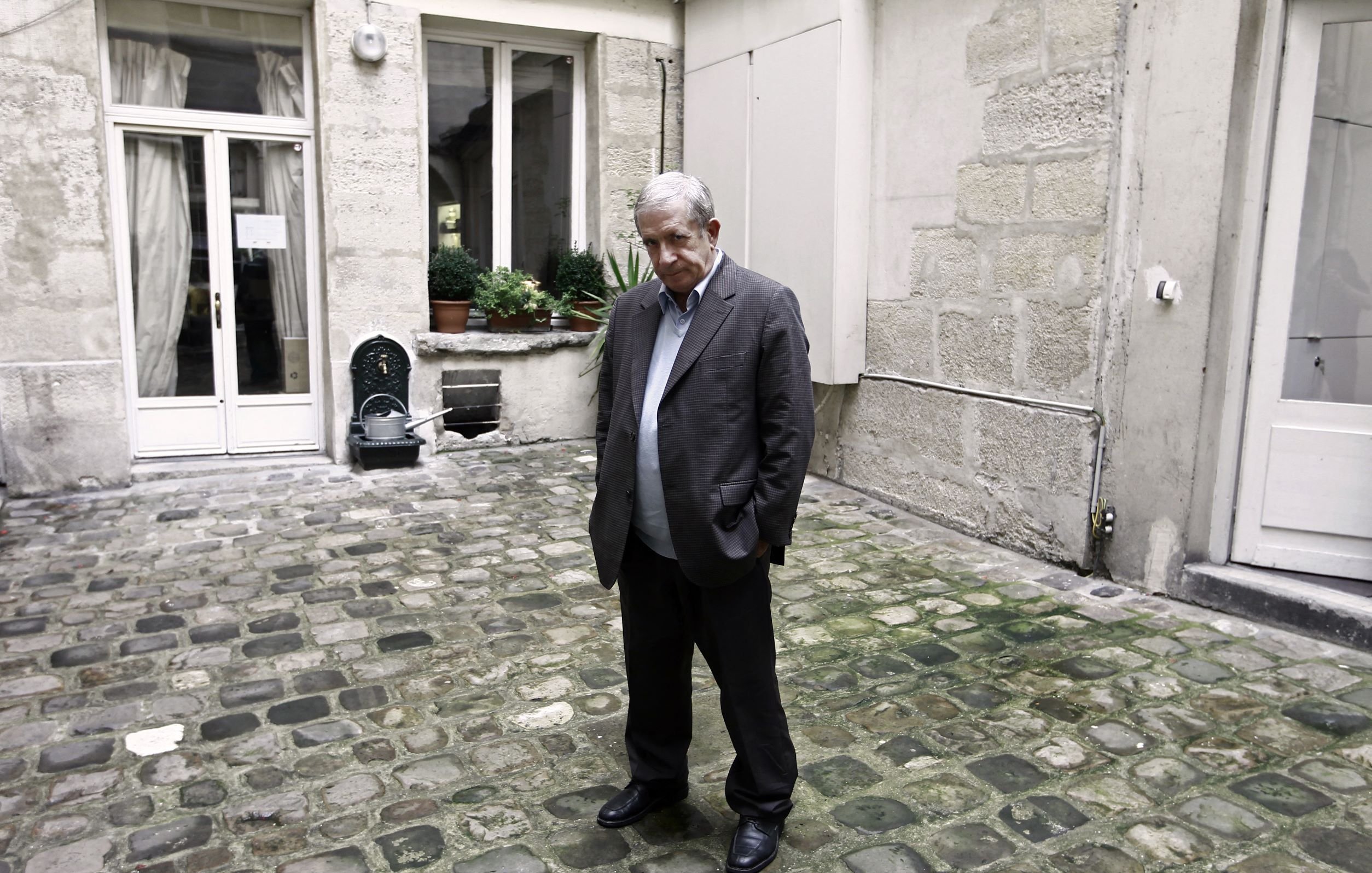 L'image montre un homme debout dans une cour pavée, visiblement pensif ou mélancolique. Il porte un costume brun et se tient les mains dans les poches. L'arrière-plan est constitué de murs en pierre et de fenêtres, avec quelques plantes en pot. L'atmosphère semble calme, mais un peu triste.