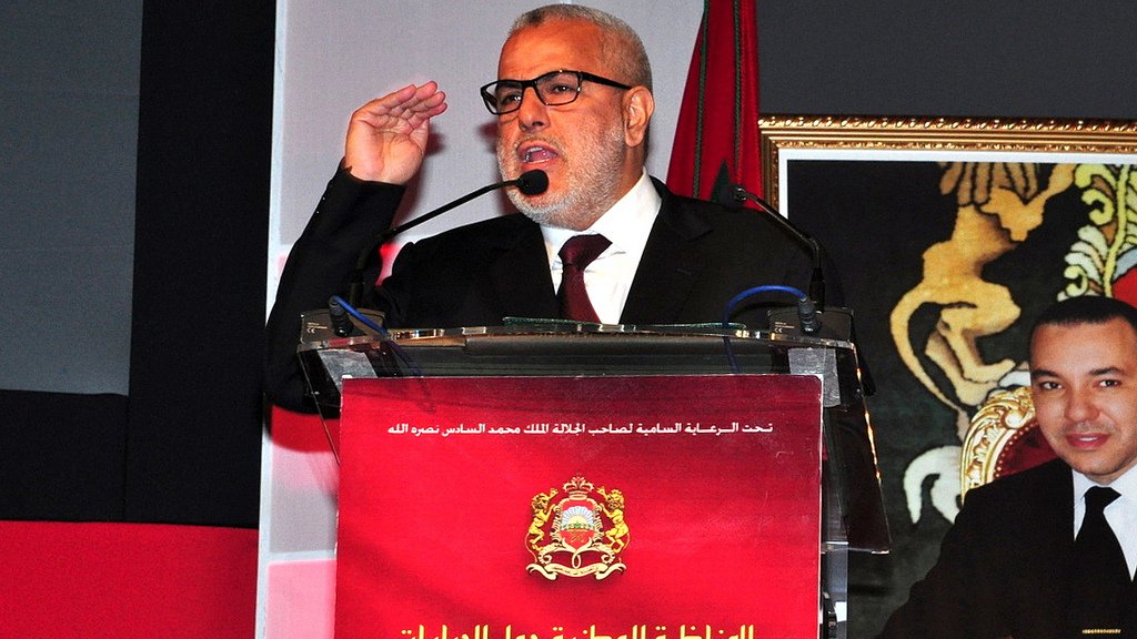 L'image montre un homme en train de s'exprimer lors d'un événement officiel. Il porte des lunettes et une veste noire, et il semble faire un geste de salutation avec sa main. Derrière lui, un panneau avec du texte en arabe et français est visible, ainsi qu'une photo d'un autre homme, probablement une figure politique. Le contexte semble être lié à des discussions sur des questions fiscales.