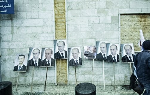 L'image montre un mur en béton avec plusieurs portraits accrochés, principalement des visages d'un homme en costume. Ces portraits sont placés sur des supports en bois. Une personne, dont on ne voit que le dos, marche à proximité. Le décor semble urbain et pourrait impliquer un contexte politique. Les expressions faciales sur les portraits sont sérieuses, reflétant une ambiance de propagande ou de soutien.