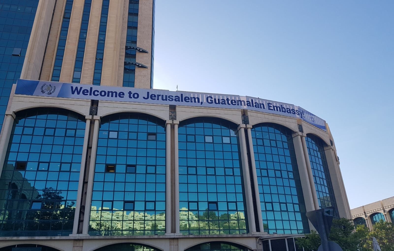 L'image montre un bâtiment moderne, probablement un immeuble administratif, avec une façade en verre réfléchissant. Au-dessus des fenêtres, une grande banderole affiche le message "Welcome to Jerusalem, Guatemalan Embassy". Le ciel est dégagé, suggérant une belle journée. Des arbres et des plantes ornent le devant du bâtiment, ajoutant une touche de verdure à l'environnement urbain.