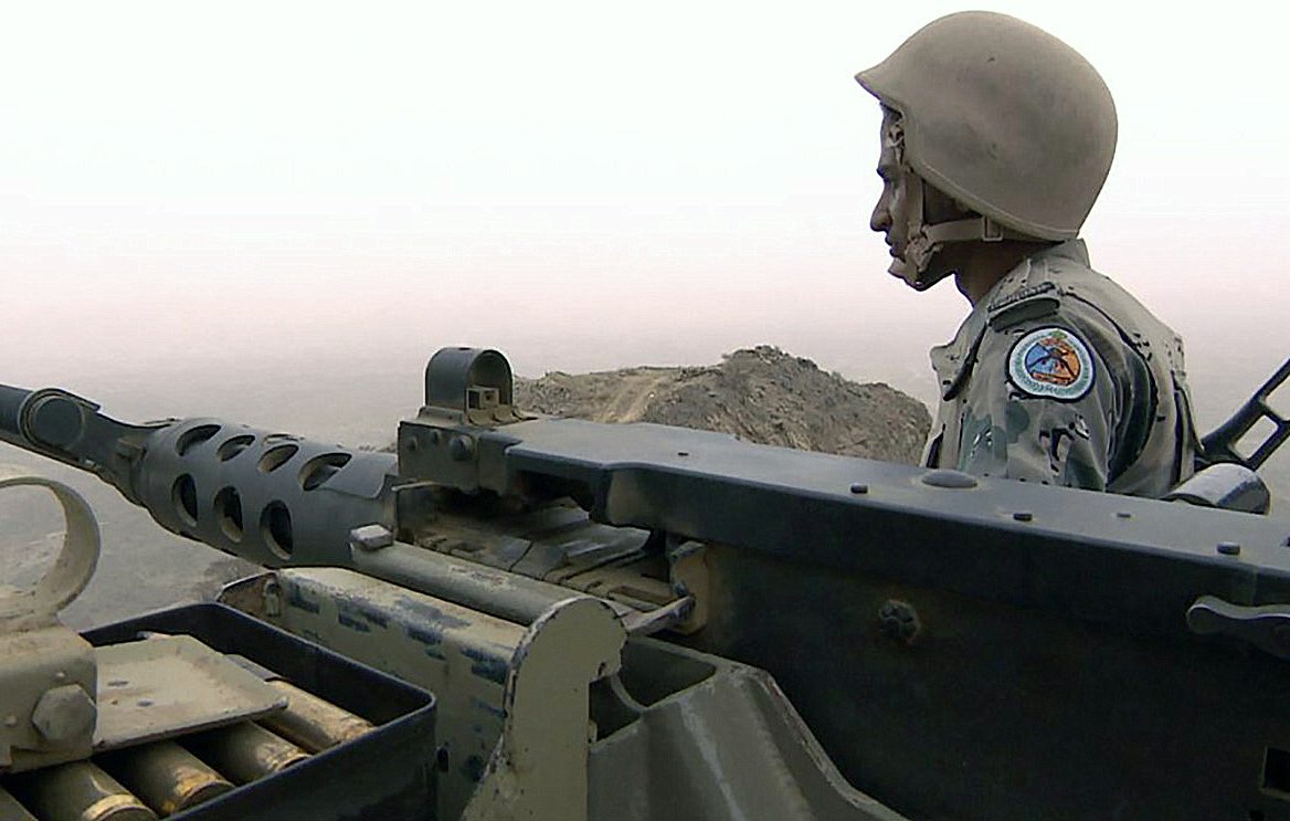 L'image montre un soldat en uniforme, portant un casque, positionné à côté d'une mitrailleuse. Il semble surveiller son environnement avec une expression concentrée. Le paysage derrière lui est brumeux, ce qui donne une atmosphère de tension et de vigilance. Le soldat est en posture d'attente, prêt à agir si nécessaire.