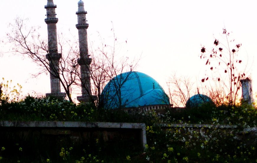 L'image montre un paysage au coucher du soleil avec deux minarets élancés se dressant vers le ciel. En arrière-plan, on peut apercevoir un grand dôme turquoise, probablement d'une mosquée, entouré de végétation. Des fleurs sauvages poussent au premier plan, ajoutant une touche de couleur et de vie à la scène. Les teintes douces du coucher de soleil créent une atmosphère paisible et tranquille.