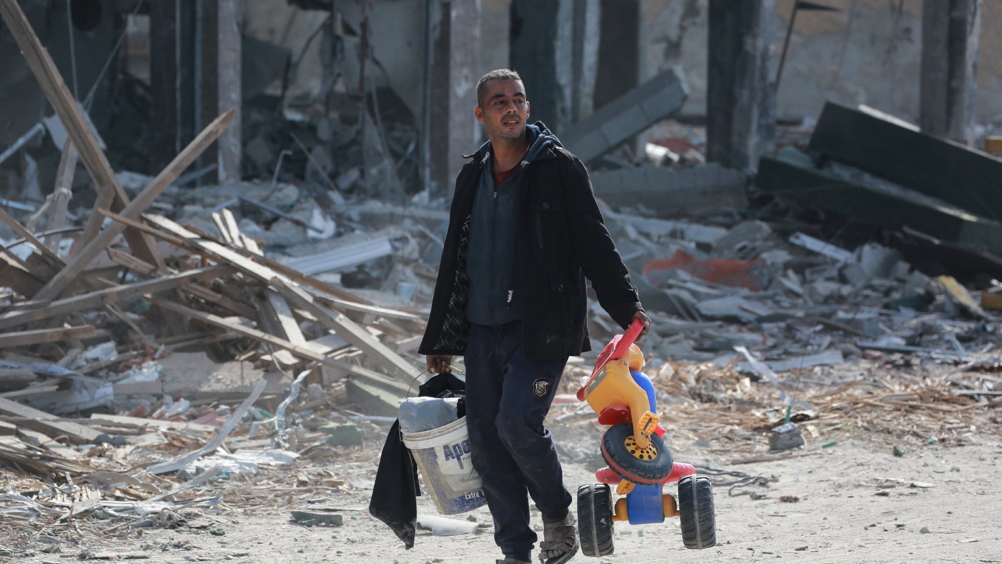 L'image montre un homme marchant dans un paysage dévasté. Il porte un seau de peinture dans sa main gauche et un jouet pour enfant, un petit véhicule à roues, dans sa main droite. L'environnement autour de lui est marqué par des débris et des ruines, suggérant un endroit qui a subi des destructions importantes. L'homme a l'air déterminé malgré la situation difficile.