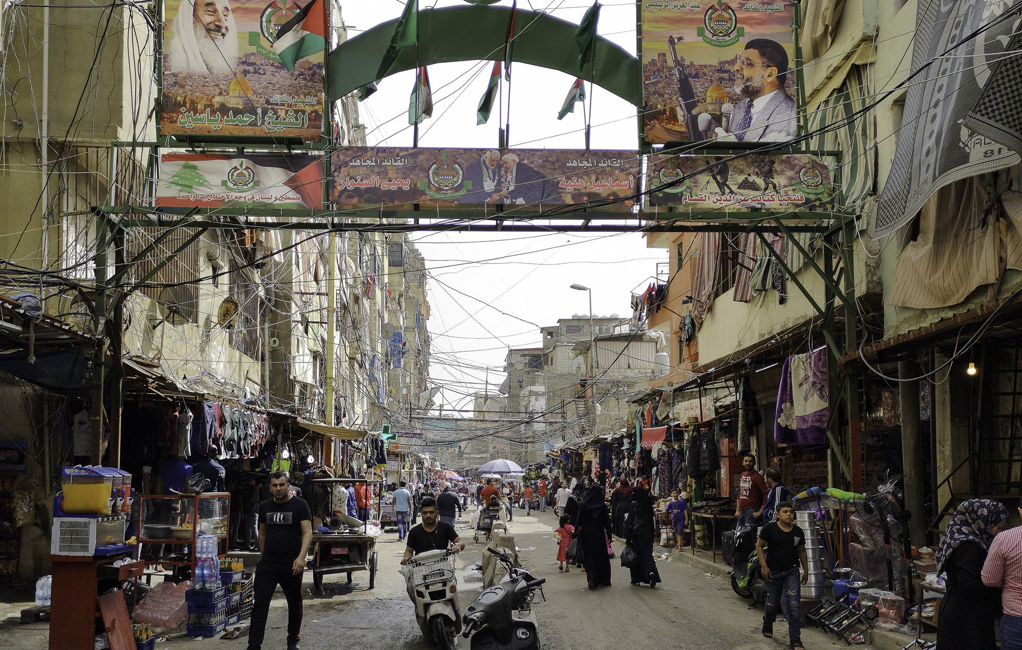 L'image montre une rue animée d'un quartier urbain. On peut y voir des bâtiments étroits et des fils électriques visibles. Des drapeaux et des affiches, probablement liés à un événement ou à une célébration, décorent l'entrée de la rue. Des personnes circulent, certaines à pied et d'autres à moto, ajoutant à l'activité de la scène. Le lieu semble être un marché ou un centre commercial, avec des étals vendant divers produits. L'atmosphère est vibrante et vivante.