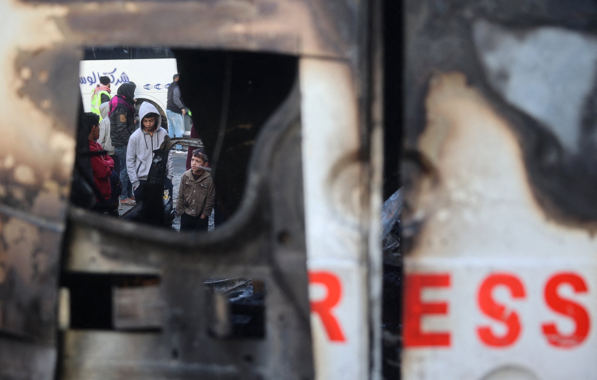 L'image montre un groupe de personnes, y compris des enfants, rassemblées dans un environnement urbain perturbé. On aperçoit une structure endommagée, peut-être un véhicule, qui présente des signes de destruction. Le reflet dans la structure suggère une atmosphère de tension ou de conflit, tandis que les visages des personnes expriment diverses émotions. L'environnement semble chaotique, soulignant une situation difficile.