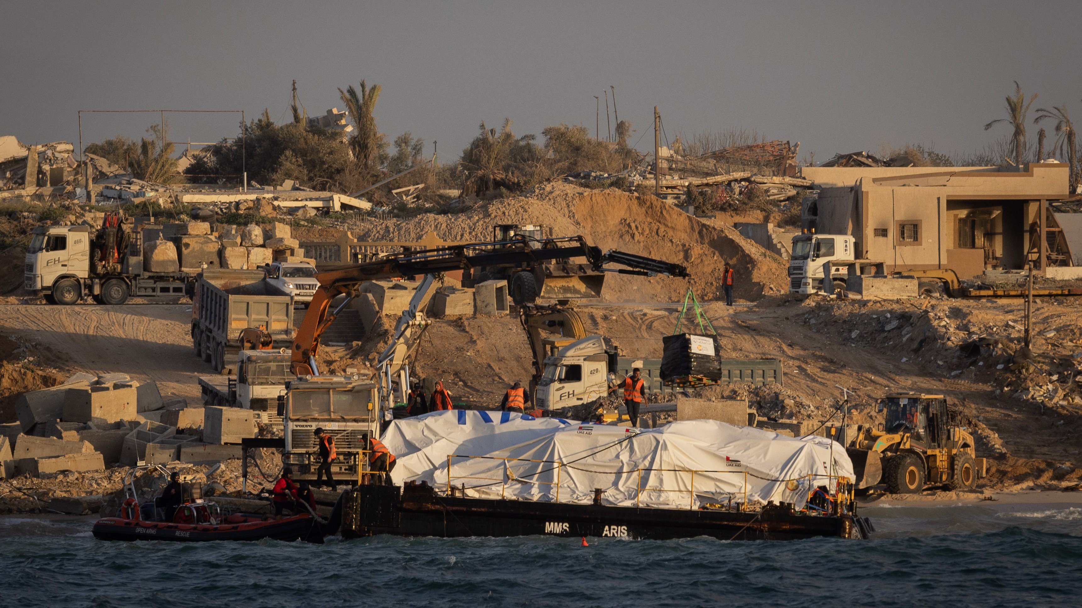 L'image montre une scène de construction ou de travaux sur une côte. On peut voir un bateau sur l'eau avec une grande bâche blanche, probablement transportant des matériaux ou des fournitures. Sur la rive, il y a des machines de construction, comme des pelleteuses et des camions, ainsi que des travailleurs en action. En arrière-plan, des bâtiments ruinés et des palmiers suggèrent un environnement en mutation, peut-être en raison de travaux de développement ou de reconstruction. L'atmosphère générale semble celle d'un chantier en cours.