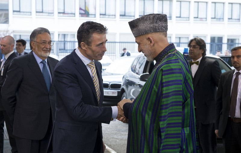 L'image montre deux hommes se serrant la main, probablement dans un cadre formel. L'un des hommes porte une tenue traditionnelle, tandis que l'autre est habillé de manière plus occidentale. À l'arrière-plan, on peut voir d'autres personnes qui semblent également habillées de façon professionnelle. L'ambiance semble être celle d'une rencontre ou d'un événement diplomatique.