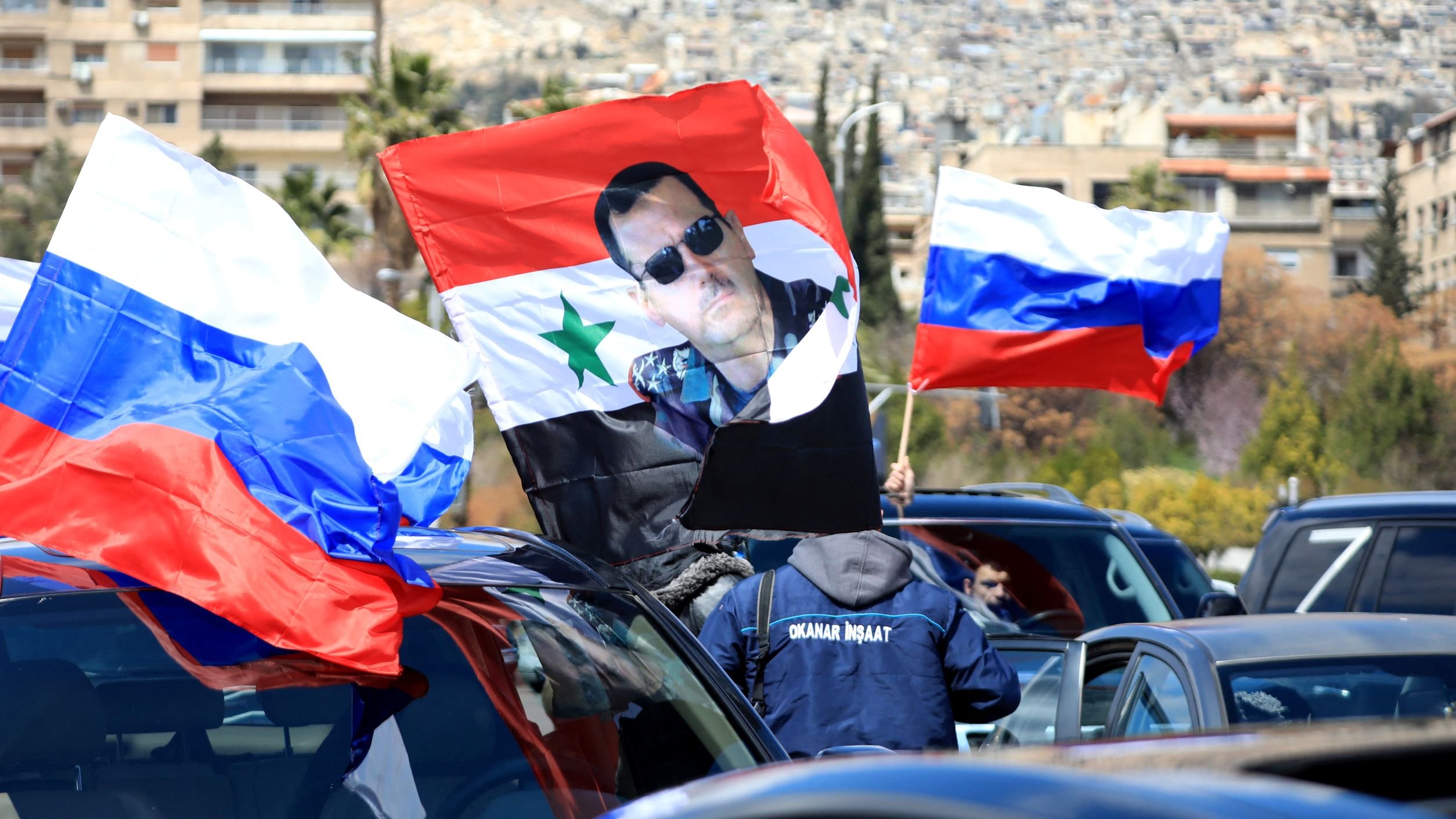 L'image montre une scène dans laquelle des personnes portent des drapeaux russes et un drapeau syrien. Au centre, on peut voir un grand drapeau sur lequel est imprimée une photo d'un homme. La scène semble se dérouler dans une ville, probablement en Syrie, avec des bâtiments en arrière-plan sur une colline. L'atmosphère suggère un rassemblement ou une manifestation, possiblement en soutien à une cause politique.
